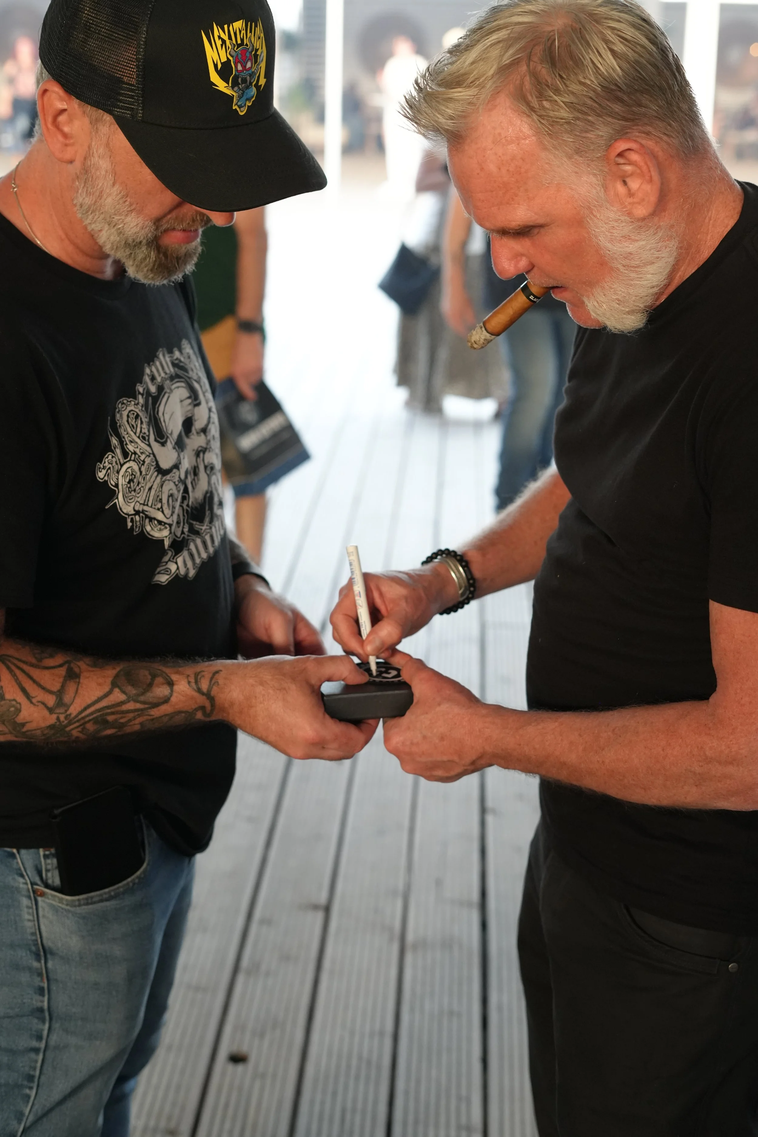 Two men with gray beards and tattoos on their arms are standing close, looking down at a black item one man is holding and signing with a white marker. One man has a cigar in his mouth and is wearing a black T-shirt. The other man is wearing a black 