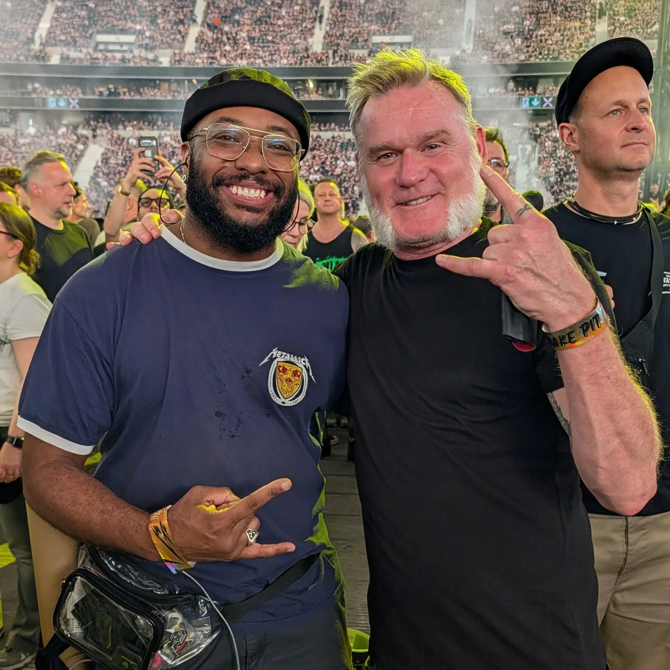 Two men posing for a photo at a crowded event. One man is wearing glasses, a black hat, and a navy blue shirt with a patch. The other man has a light-colored beard, is pointing at his head, and is wearing a black shirt. The background shows many people attended the event, some taking photos, with a large crowd in a stadium or arena.