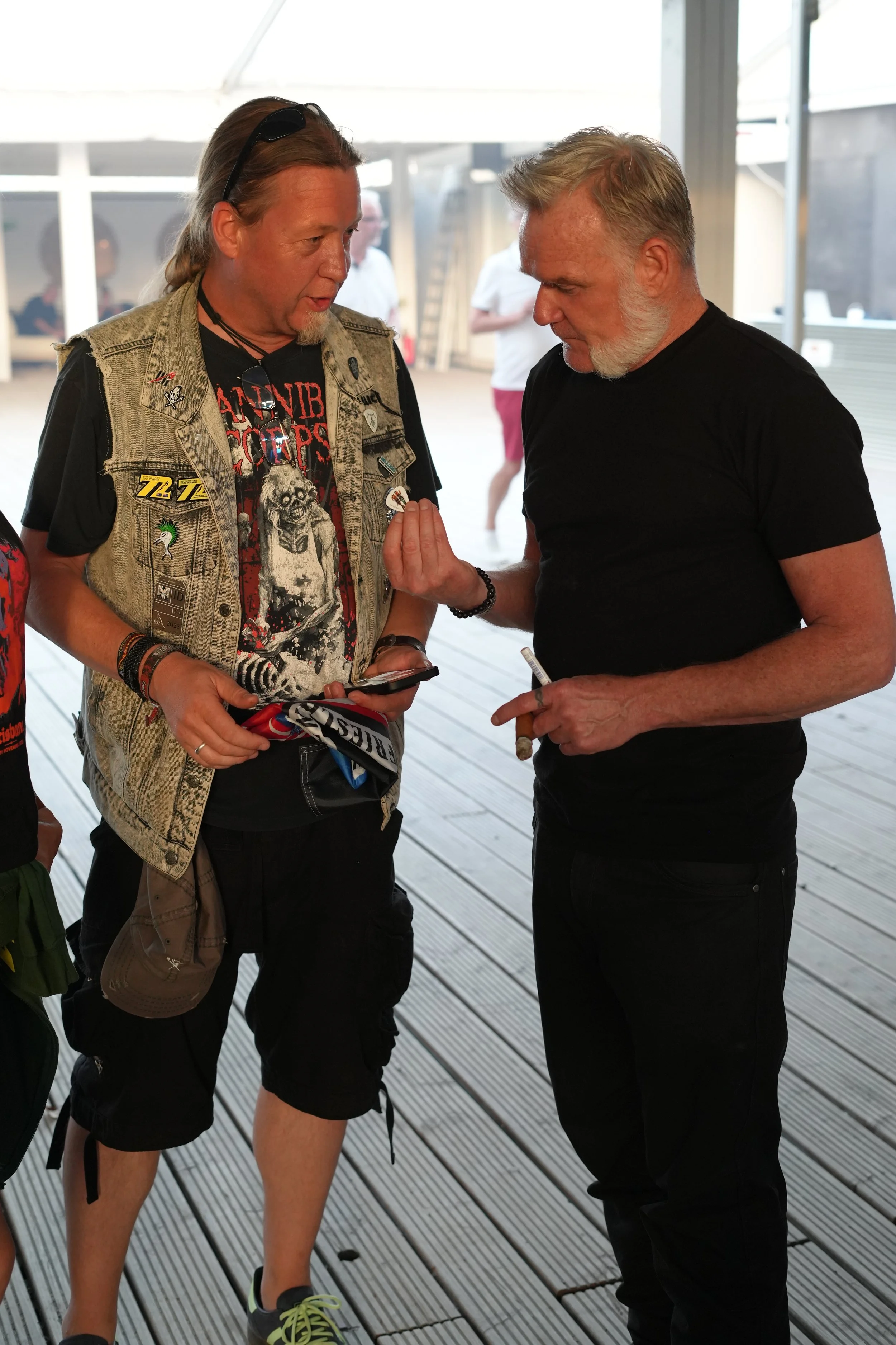 Two middle-aged men with gray hair having a conversation indoors. The man on the left is wearing a denim vest with various patches, a graphic t-shirt, and sunglasses hanging from his neck. The man on the right is dressed in a black t-shirt and is hol