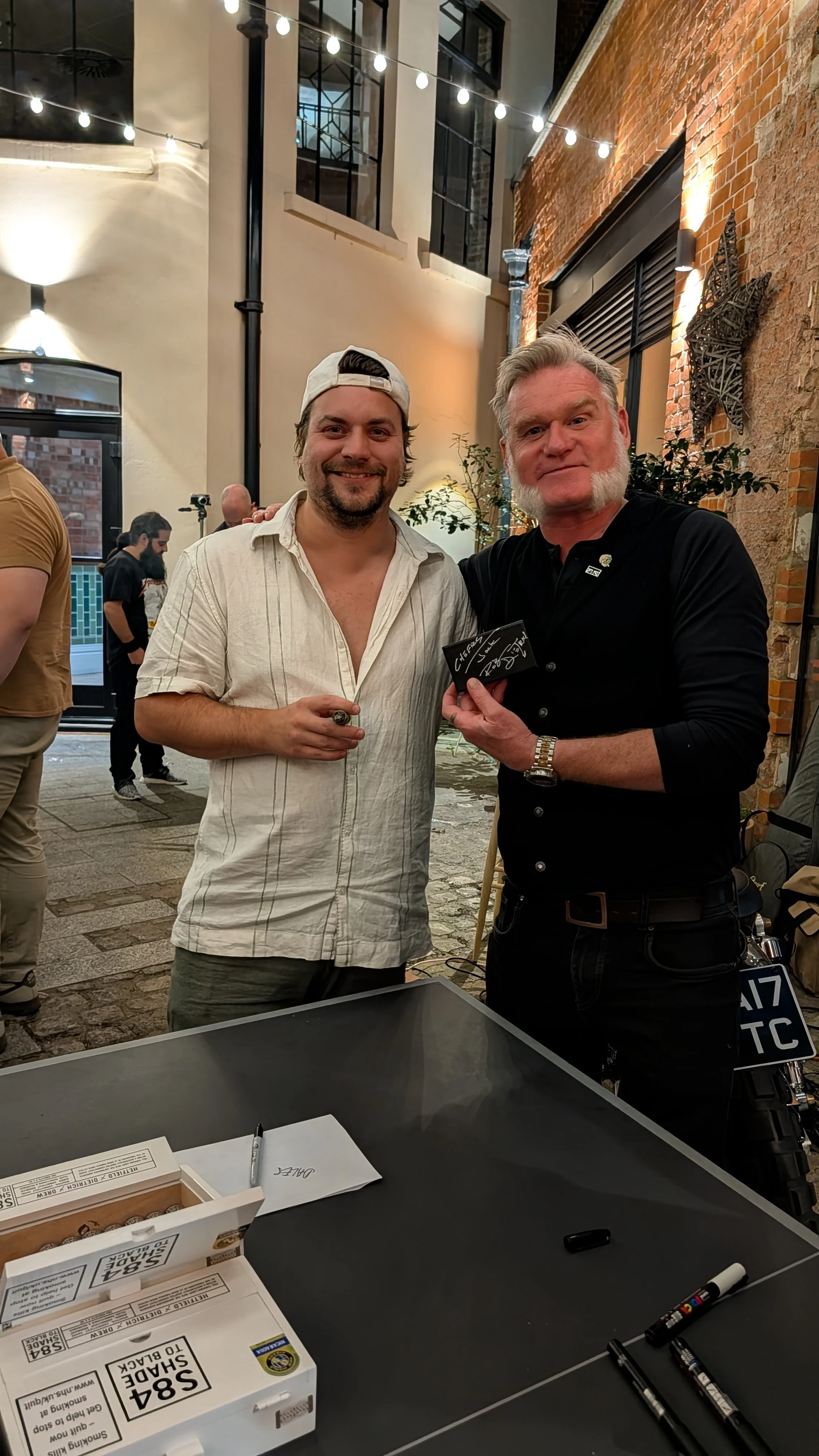 Two men standing inside a well-lit industrial-style venue, one making a peace sign and the other holding a small blackboard with signatures. There are other people in the background, and the scene includes brick and white walls, hanging string lights