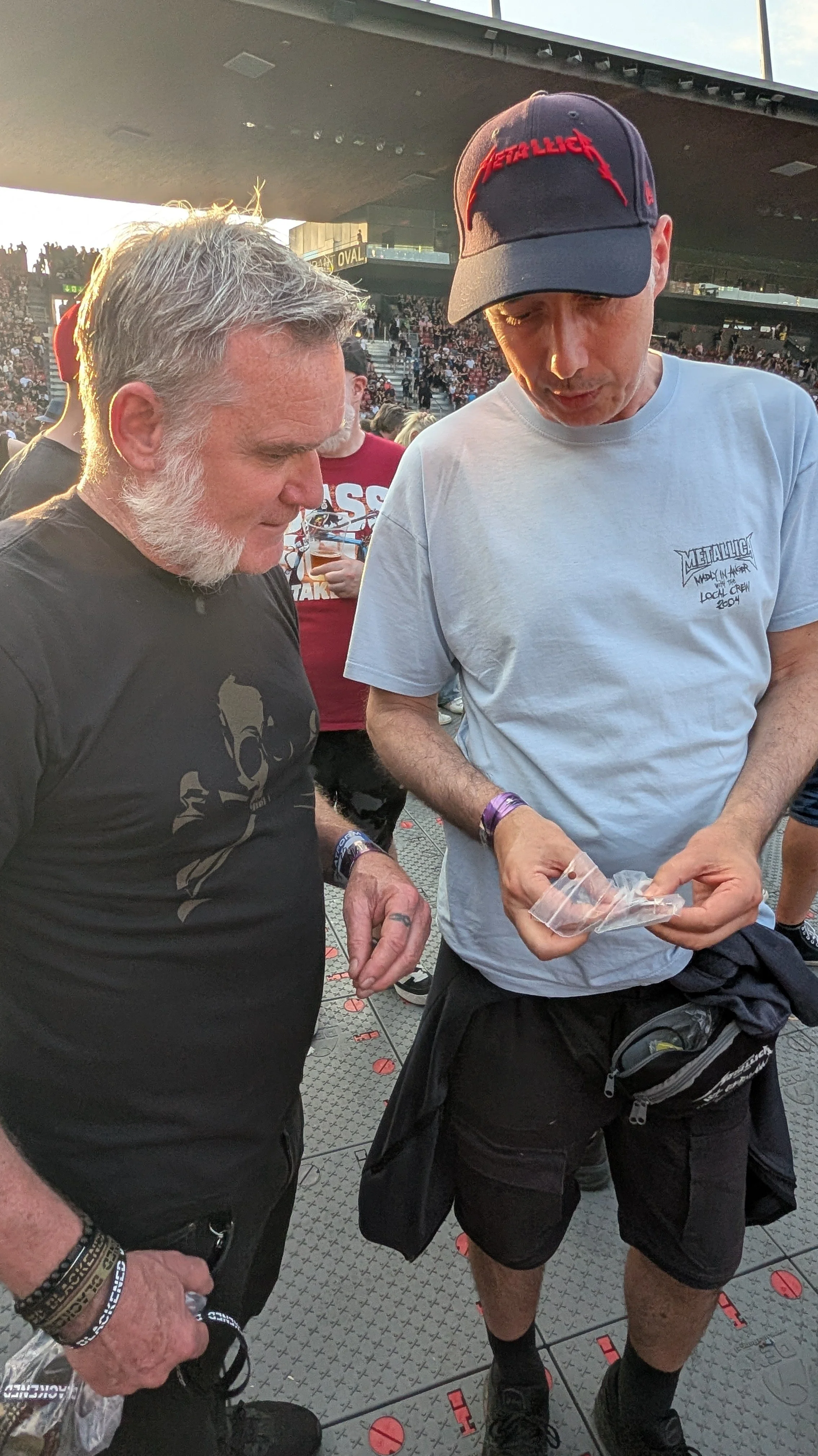 Two men standing close together at a Metallica concert looking down at guitar picks in their hands. 