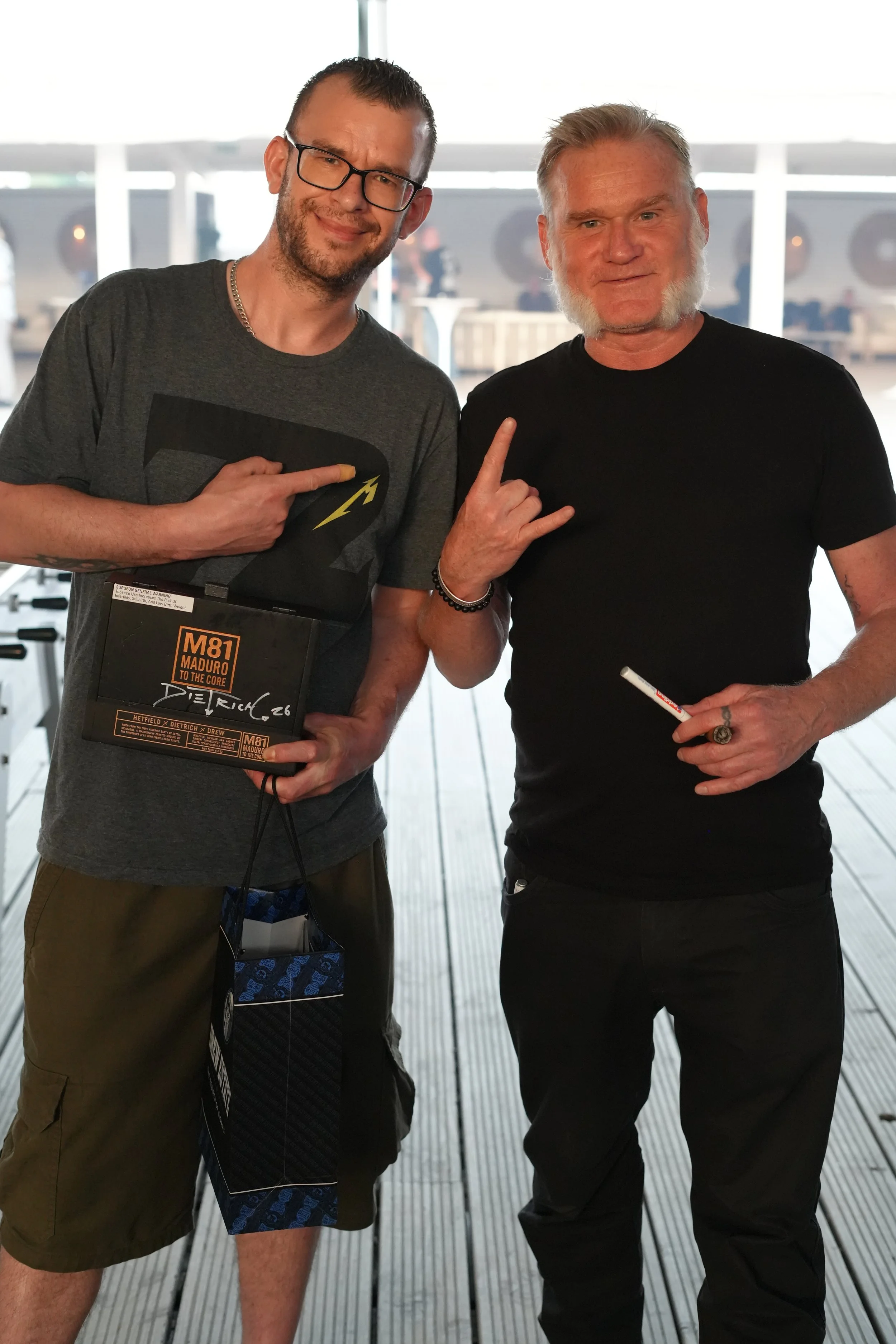 Two men standing together indoors, both making the 'rock on' hand gesture. The man on the left is wearing glasses, a gray t-shirt with a superhero logo, and khaki shorts, holding a book. The man on the right has gray hair and a beard, wearing a black
