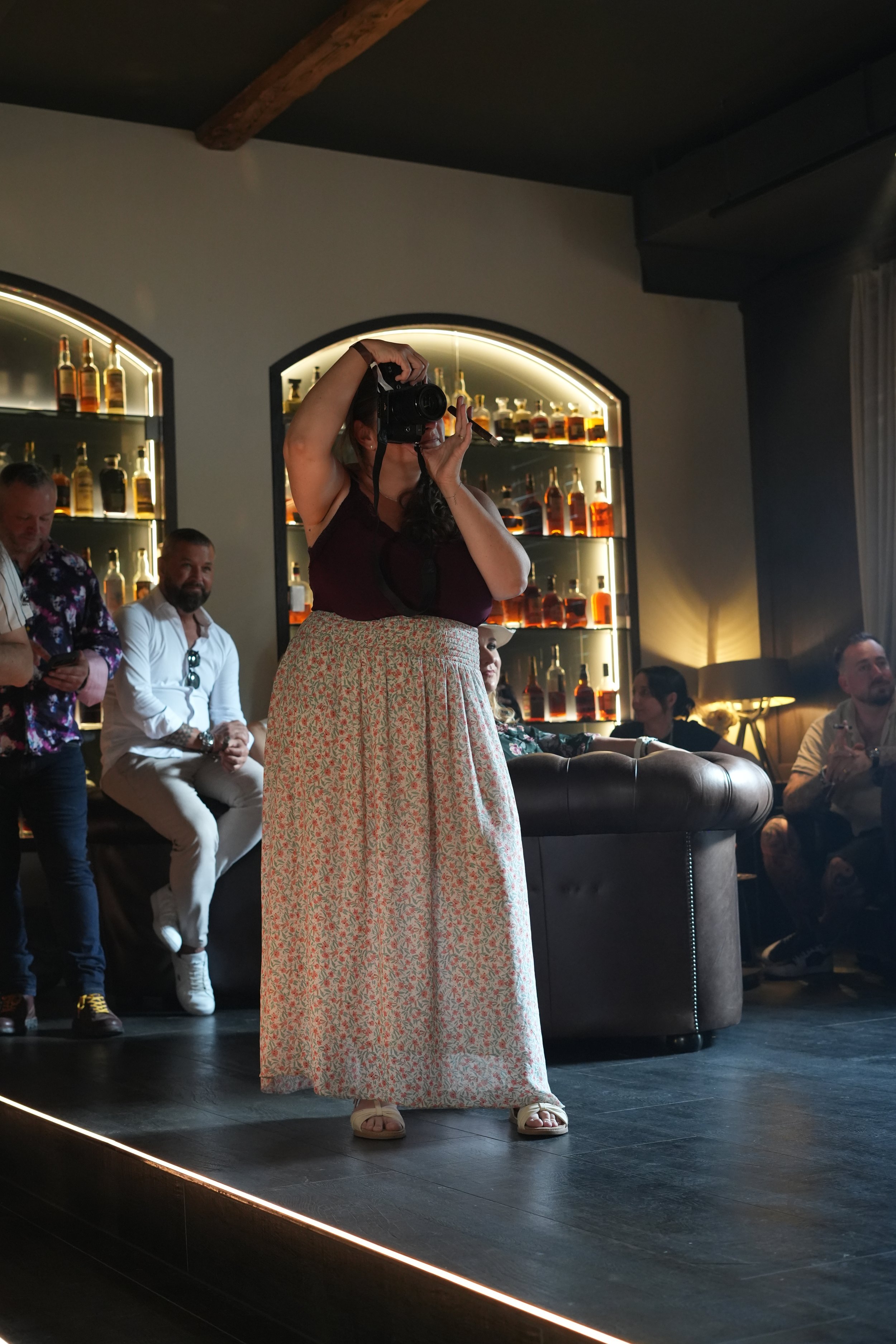 A woman standing on a stage taking a photo with a camera, with suspenders, in a room with shelves of liquor bottles and other people sitting and standing nearby.