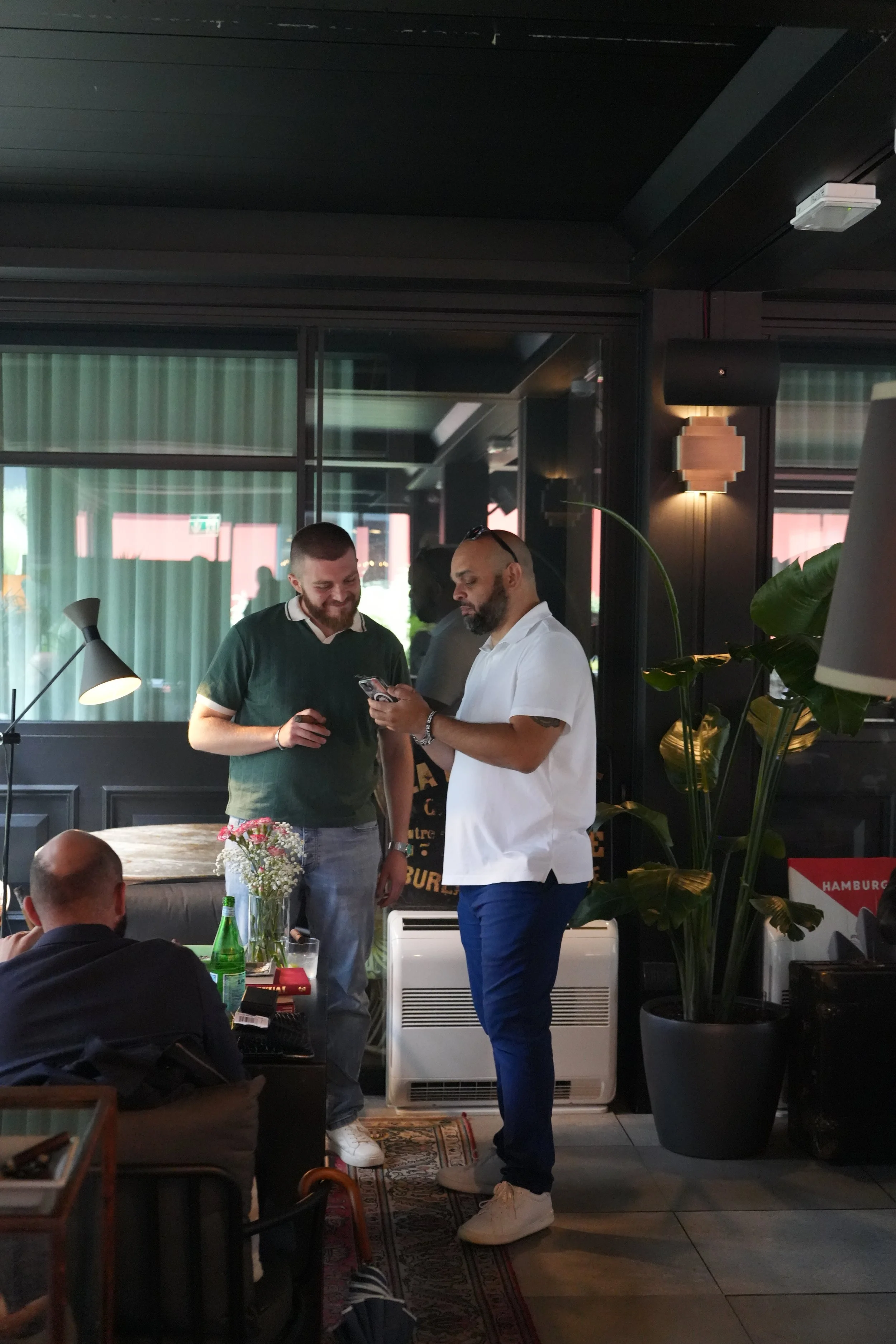 Two men are standing and looking at a phone inside a restaurant or cafe. One is wearing a black shirt and the other a white shirt. There are other people, tables with drinks, a plant, and decorative lighting in the background.