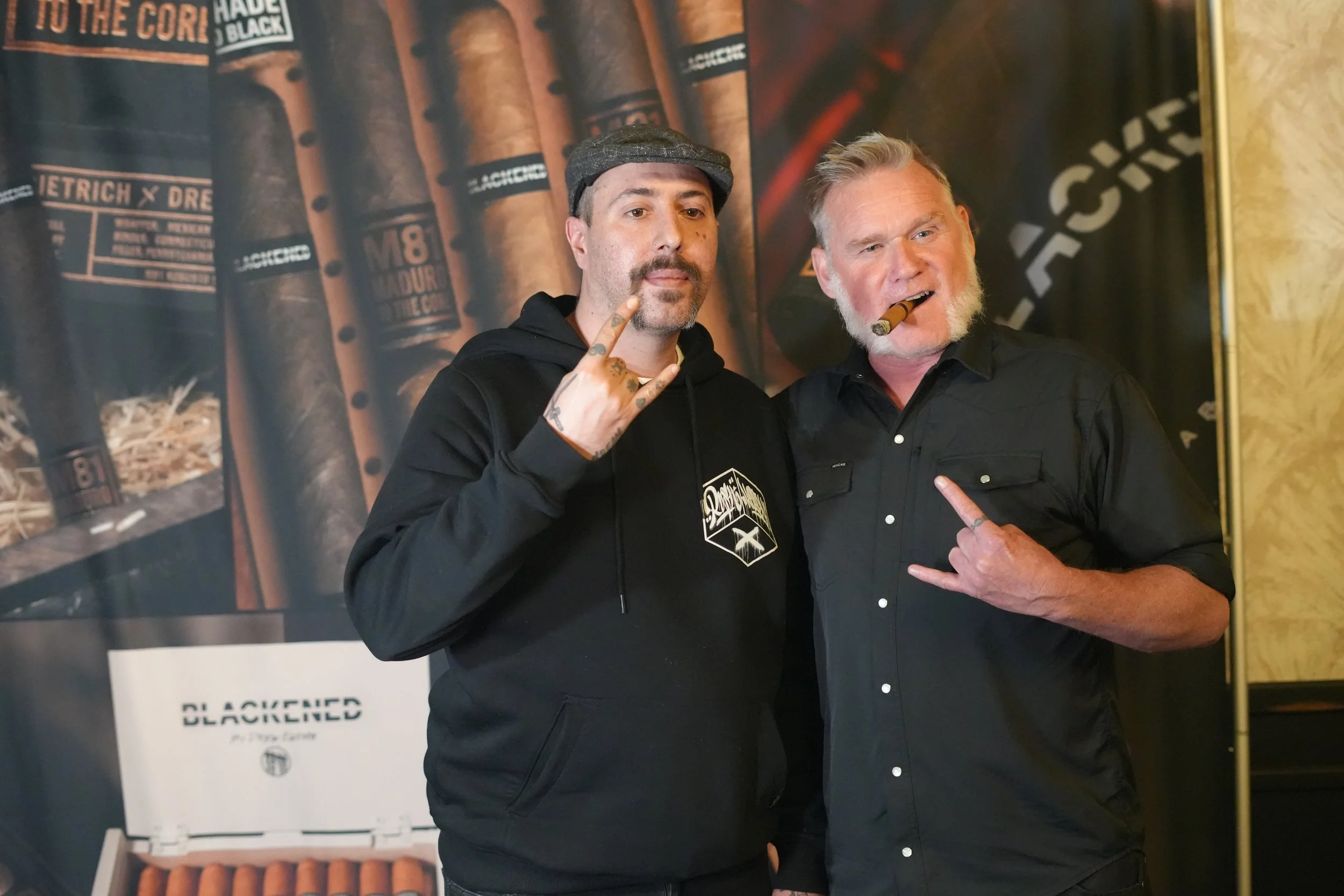 Two men standing side by side, both making a rock 'n' roll hand gesture and holding cigars in their mouths. The man on the left is wearing a gray flat cap and a black hoodie, and has tattoos on his fingers. The man on the right has short, gray hair a