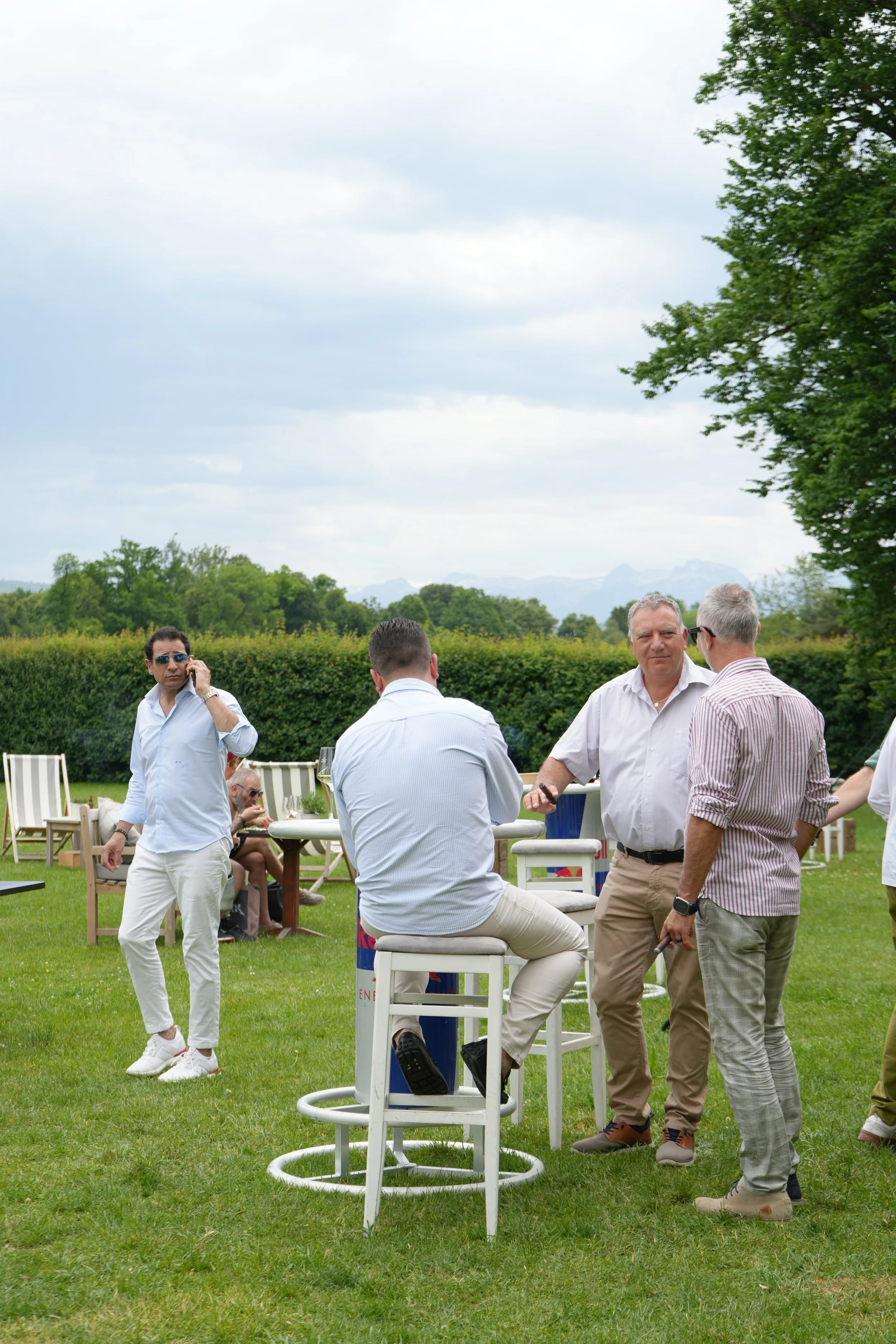 A group of men socializing outdoors on a grassy area with chairs and a bar table, with a hedge, trees, and mountains in the background on a cloudy day.