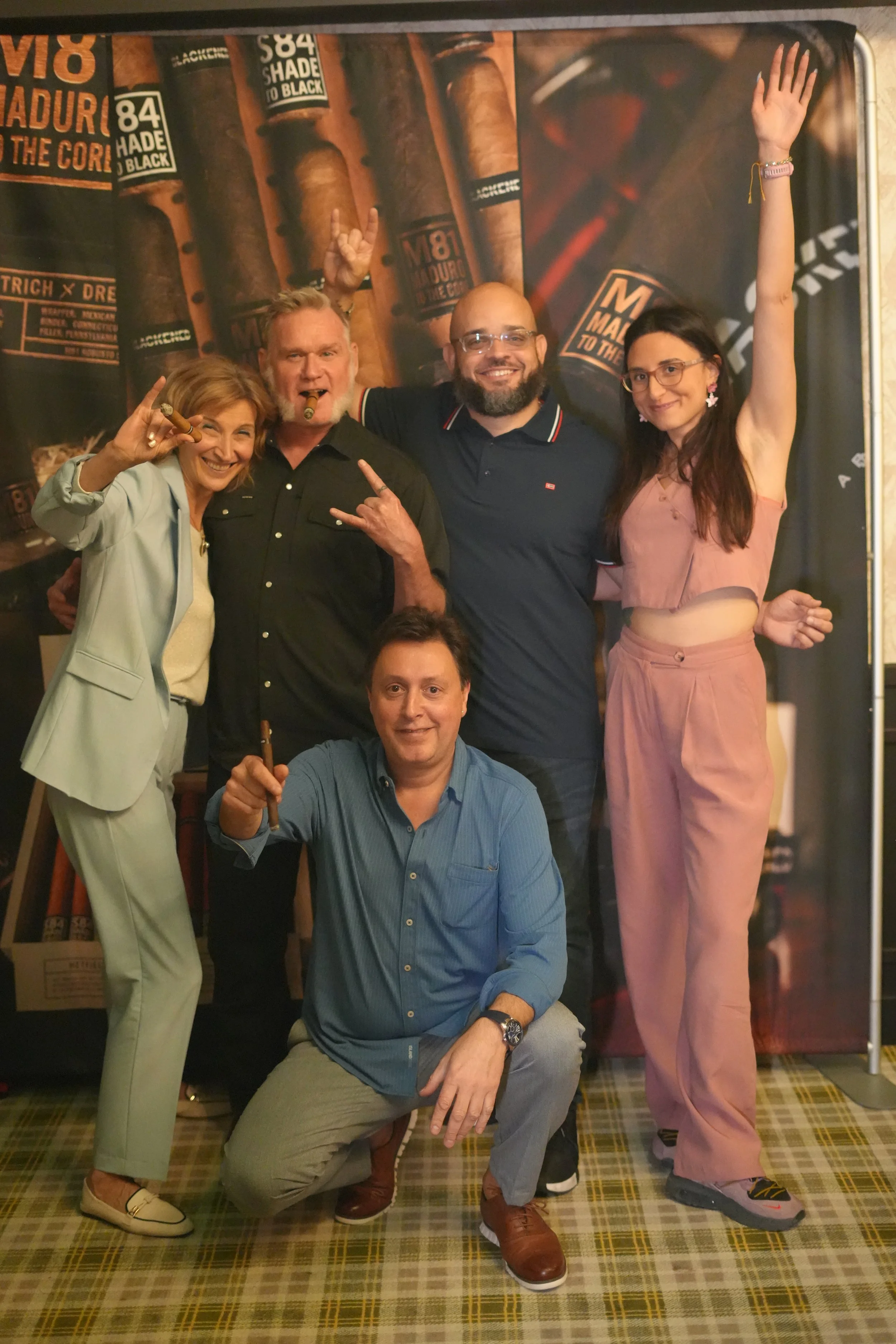 Four people posing in front of a cigar-themed backdrop, some holding cigars, with one person giving the 'rock on' hand sign, all smiling.