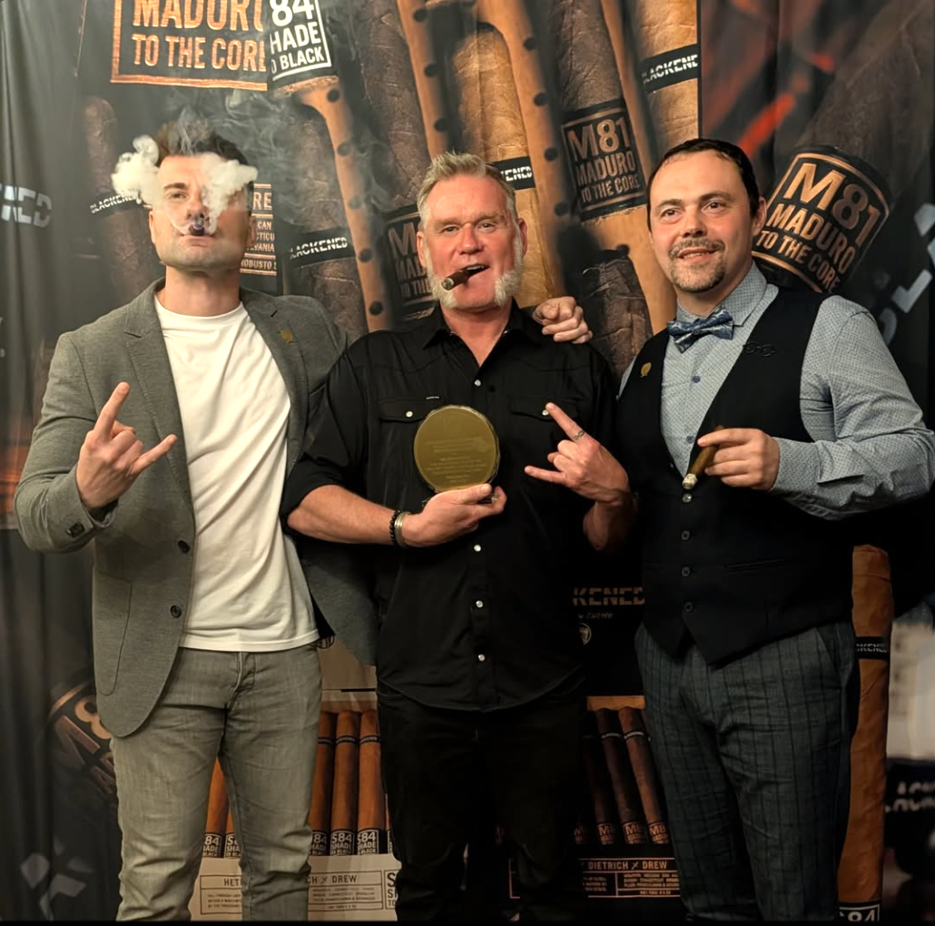 Three men posing at a cigar event, one holding a cigar and a cigar award. The man on the left is making a rock hand gesture and smoking a cigar with smoke visible. The man in the middle has a cigar in his mouth, a beard, and is holding a cigar award.