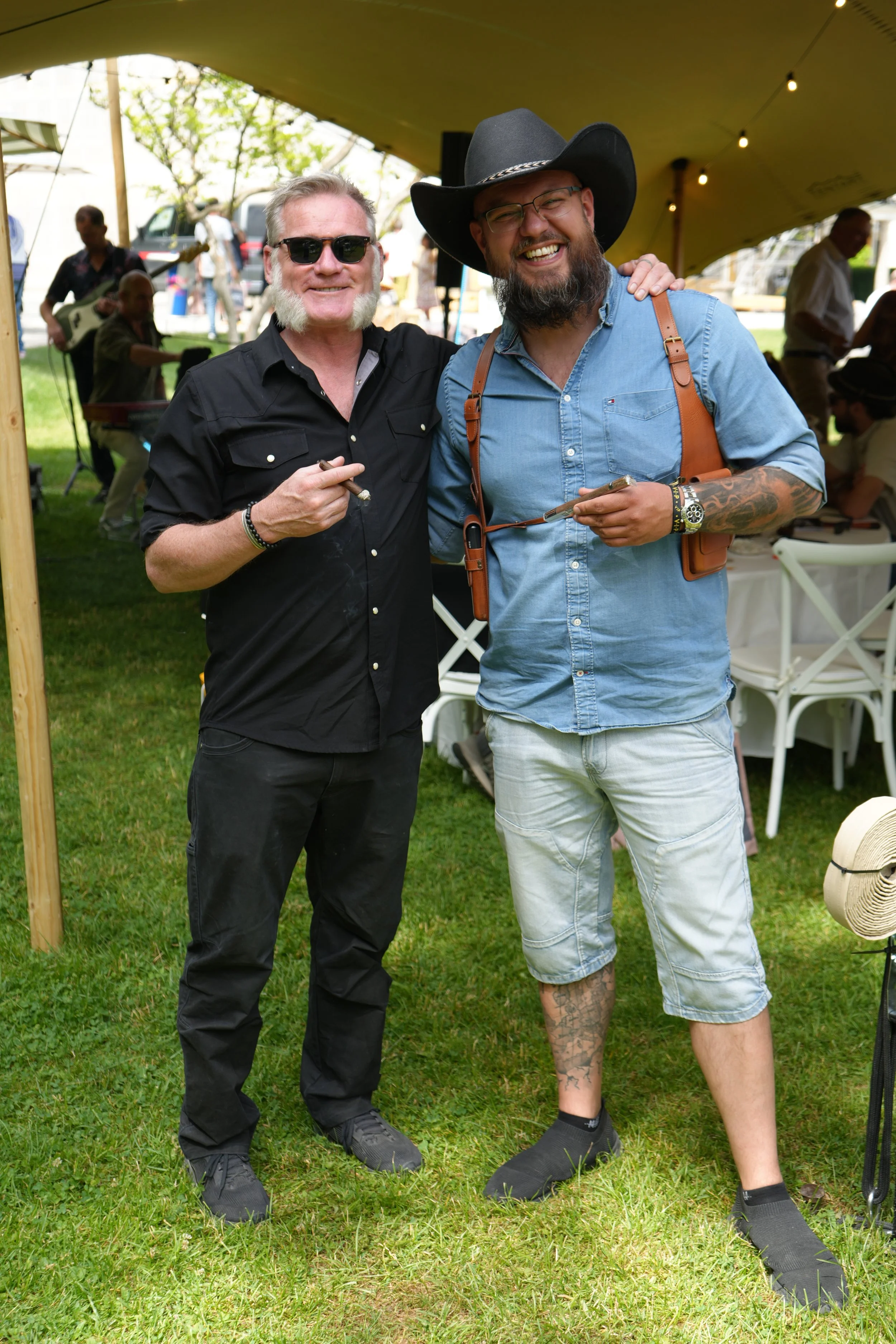 Two men standing outdoors in a grassy area under a yellow canopy, smiling and posing for a photo. The man on the left has short gray hair, wears black sunglasses, a black button-up shirt, and black pants. The man on the right is wearing a black cowbo