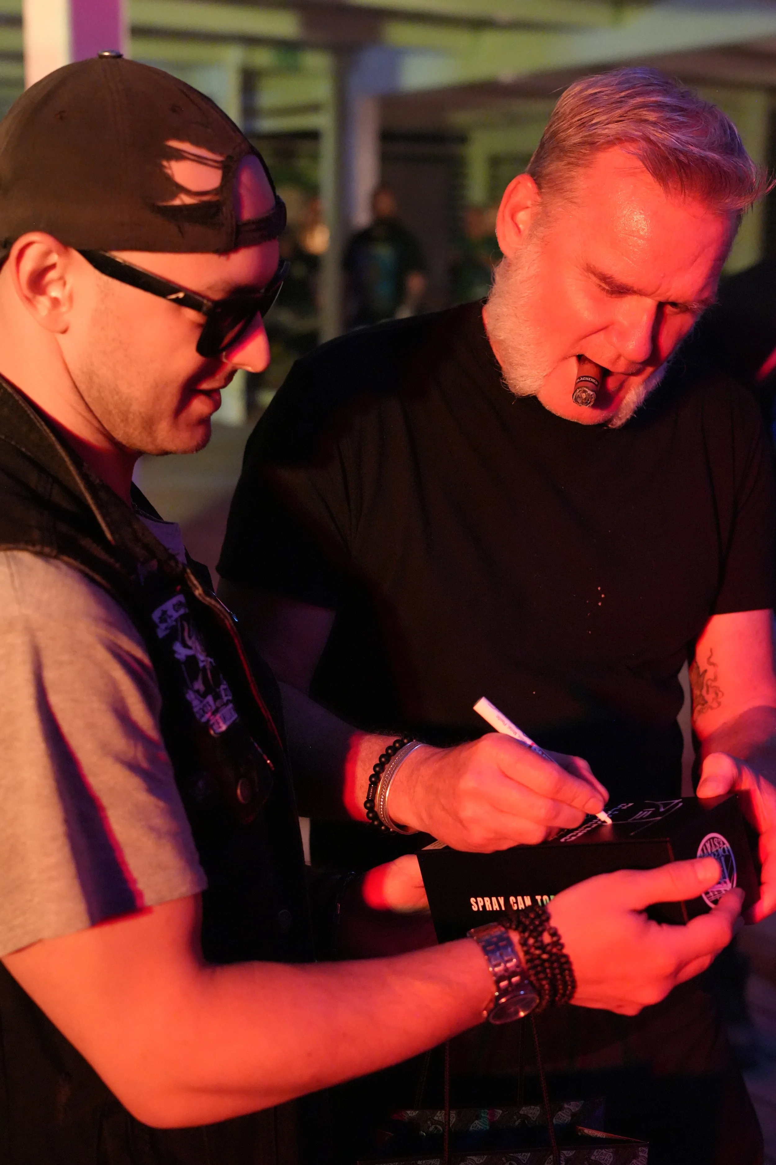 Two men at a social event, one with a cigar in his mouth, looking at a signed poster or card. One wears sunglasses and a black cap with ripped details, the other has gray hair and a beard. The environment is lit with red and purple lighting.
