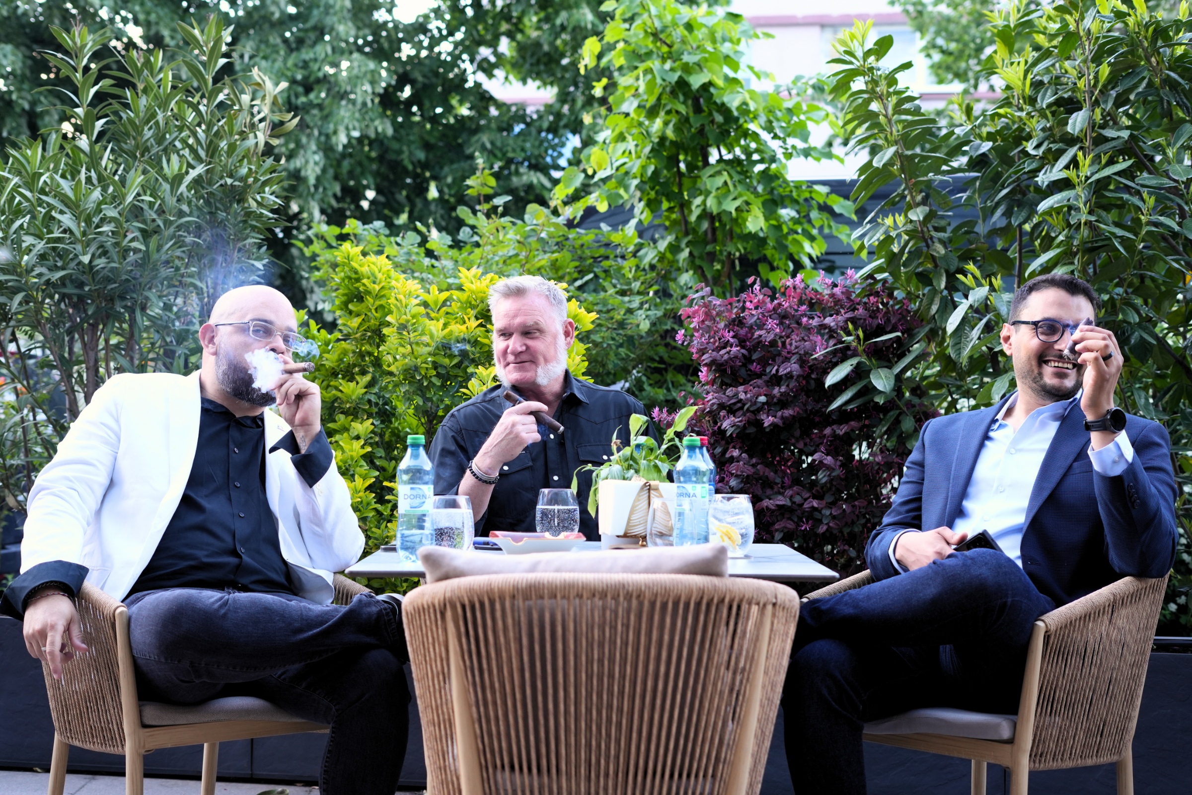Three men sitting at an outdoor table surrounded by lush green plants, drinking and smiling during a social gathering.