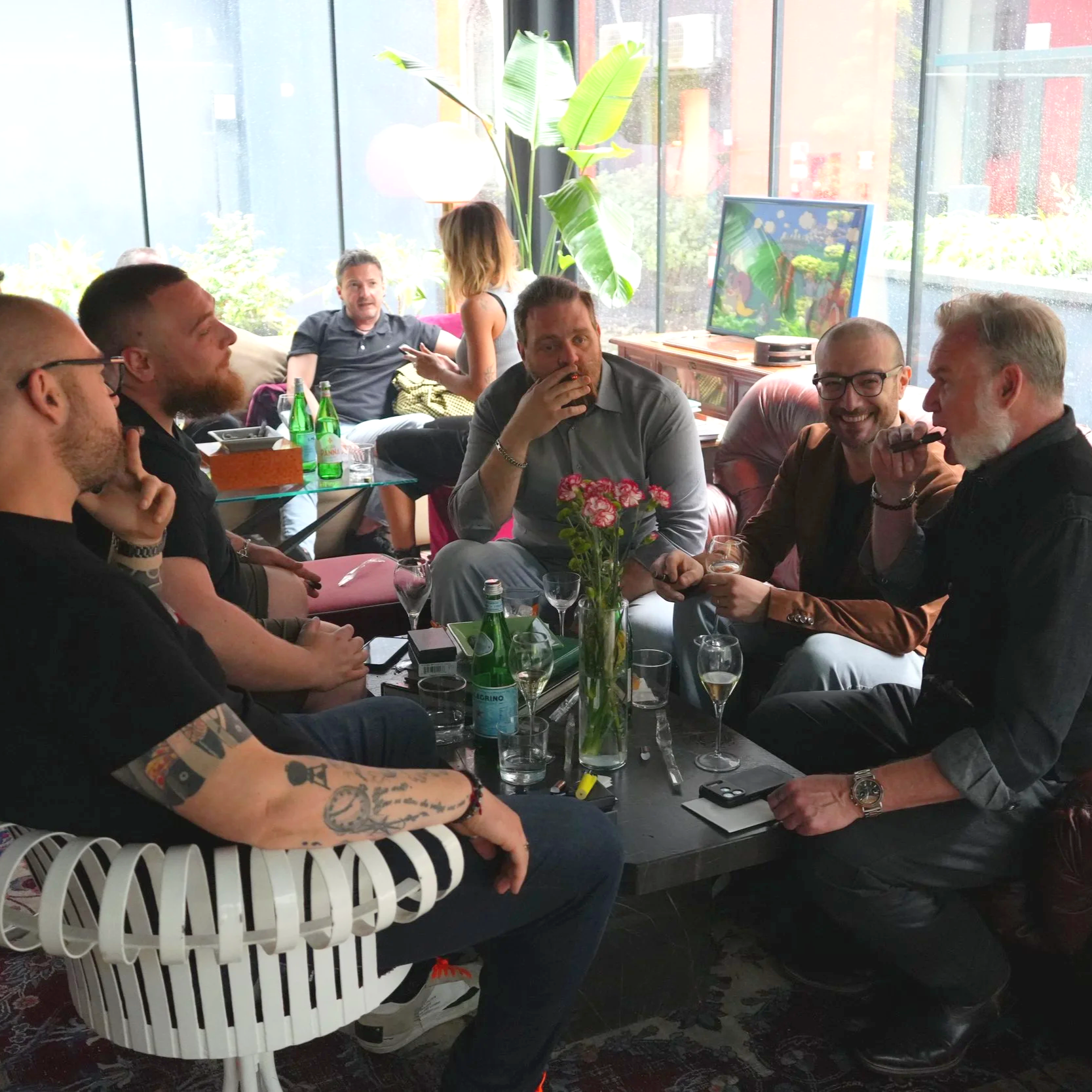 Group of people gathered in a brightly lit room with large windows, sitting around a table with drinks and a flower centerpiece, engaging in conversation.