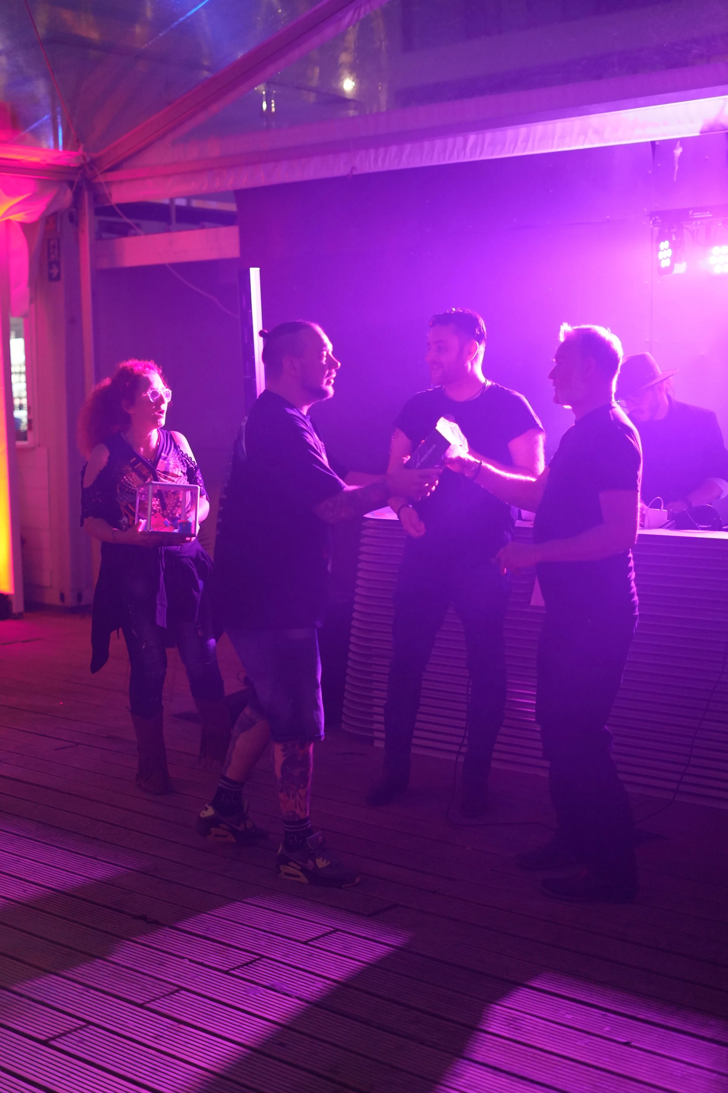 Five people talking and holding items under purple and pink lighting in a room with wood flooring.