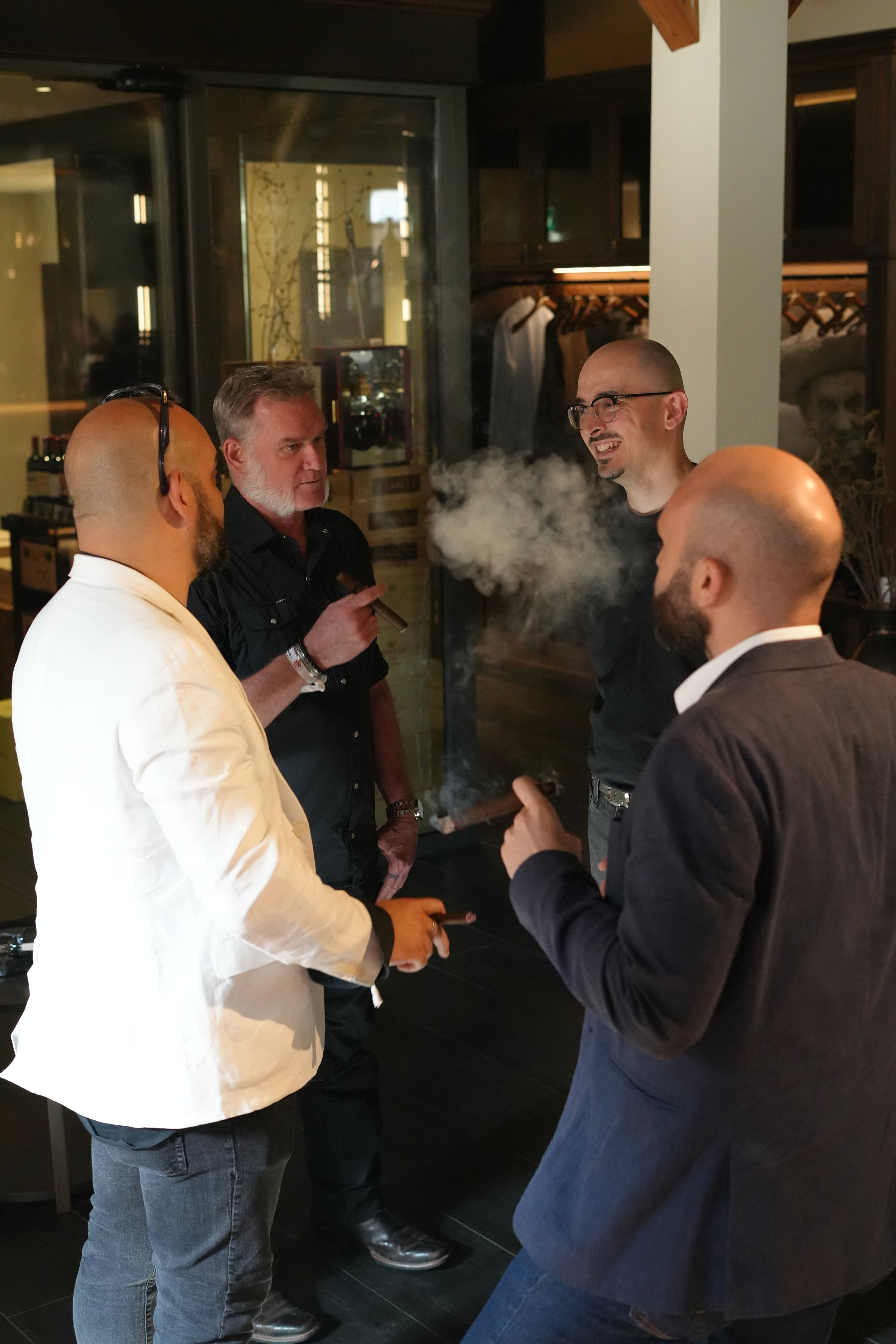 Four men gathered in a restaurant or bar, having a conversation. One man with a beard and glasses is smiling, while another man with a beard and a white blazer is holding a cigar. The other two men have short hair, one with glasses and a black shirt,