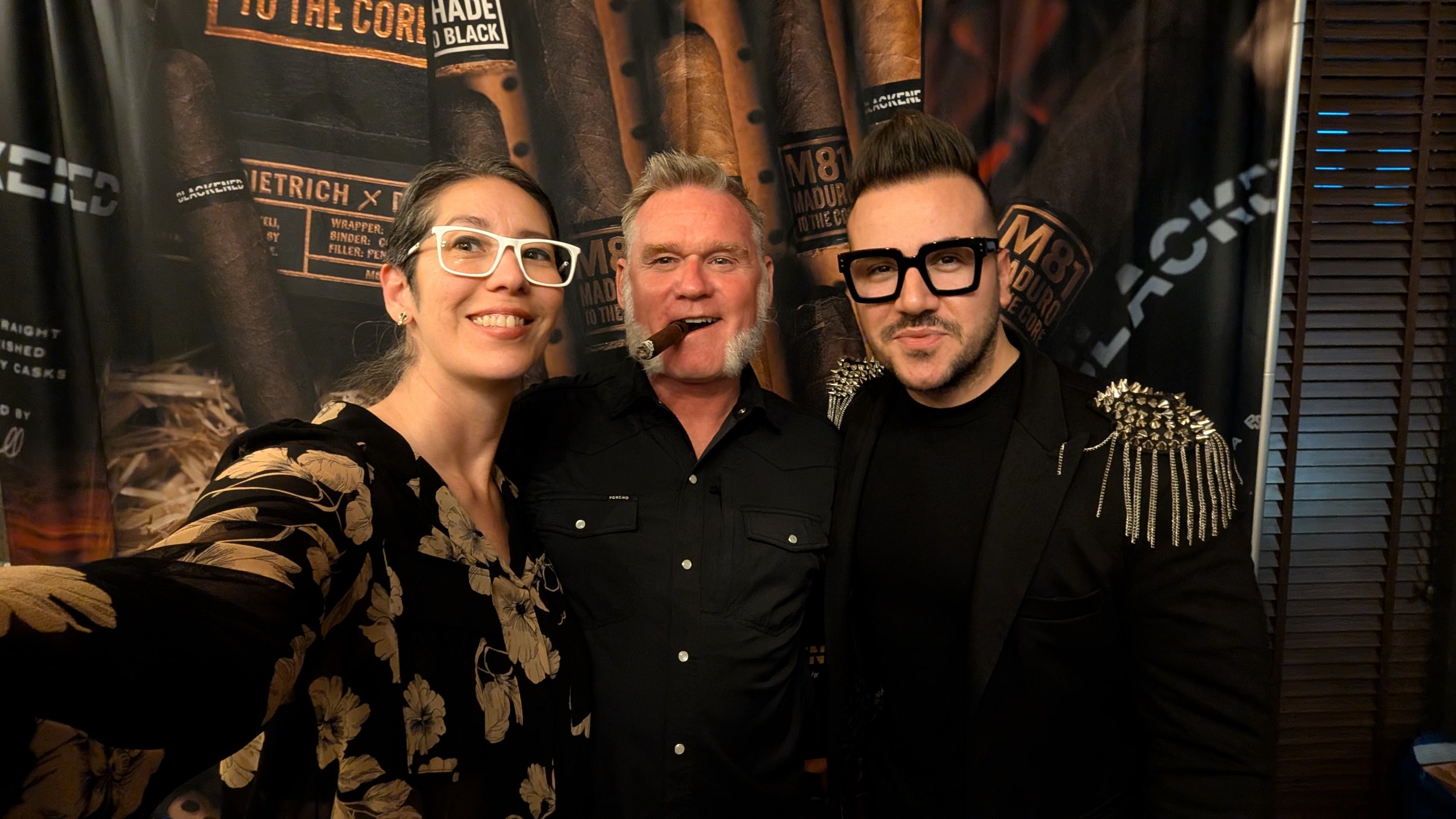 Three people posing together at an event, standing in front of a large cigar-themed backdrop.