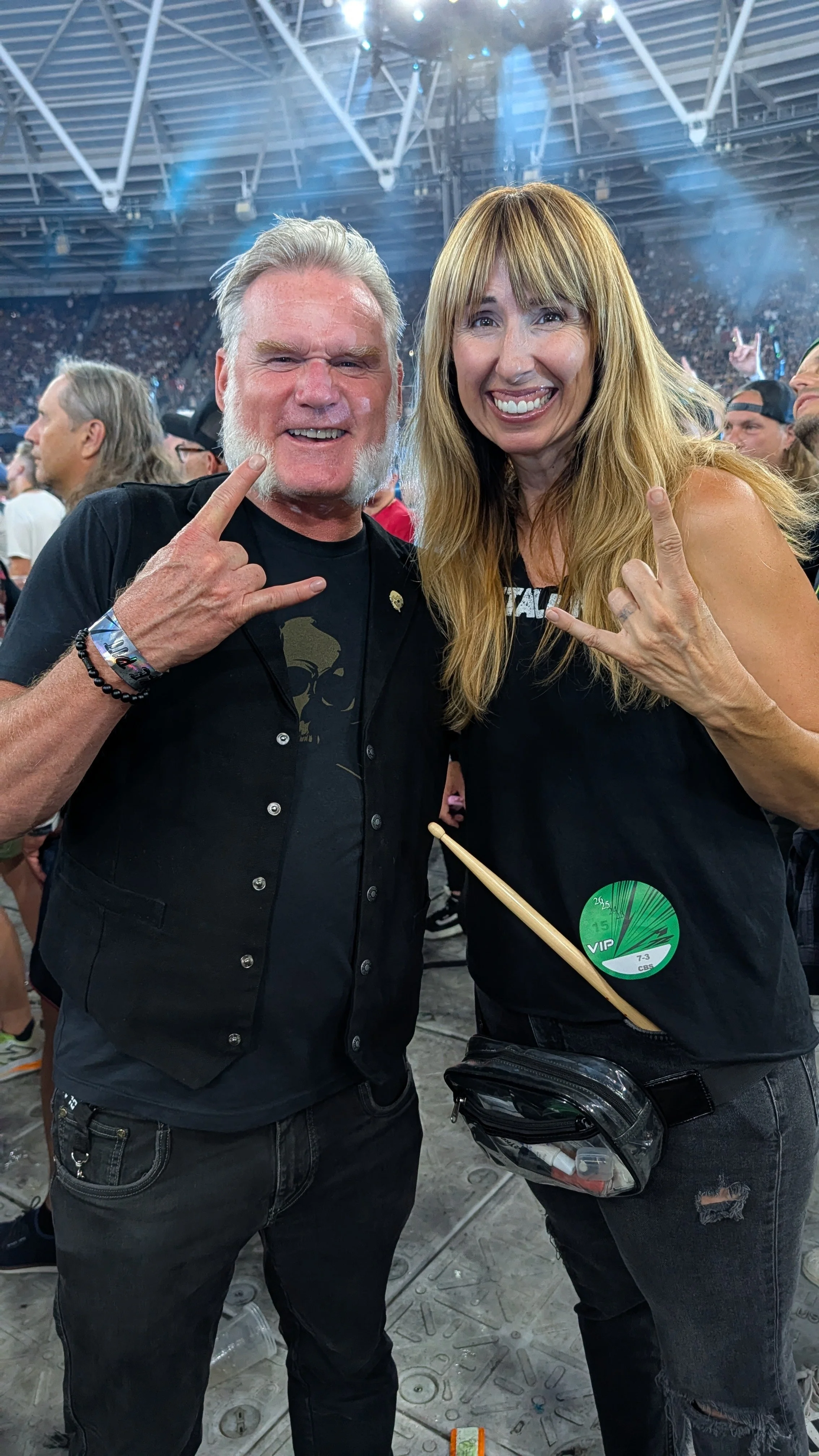 Two people posing at a concert, both making rock signs with their hands. The man has gray hair and a beard, wearing a black vest and black jeans. The woman has long blond hair, wearing a black top with a green VIP pass attached, and distressed black 