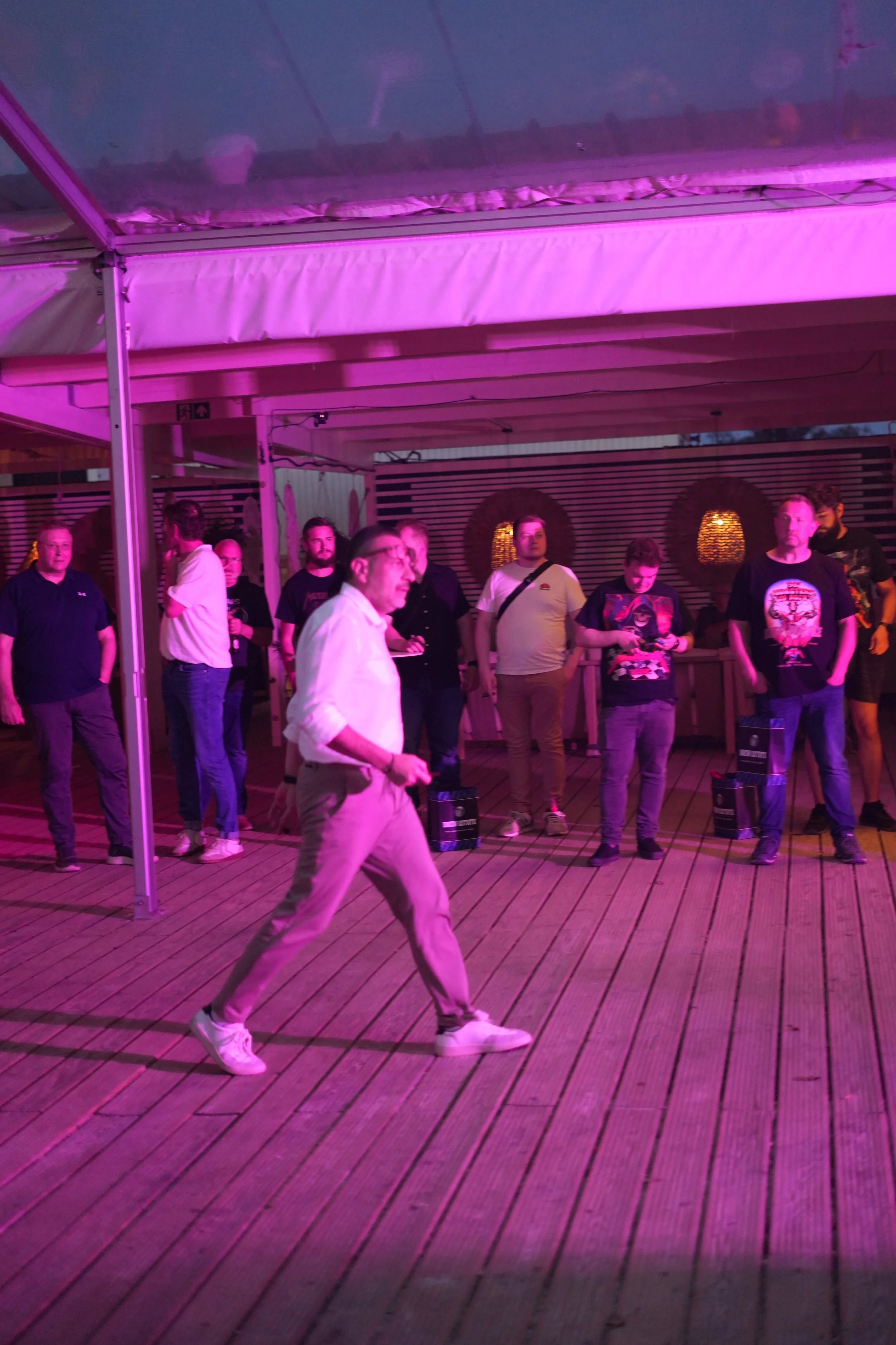 A group of people standing on a wooden deck under purple lighting, with some looking at their phones and others observing.