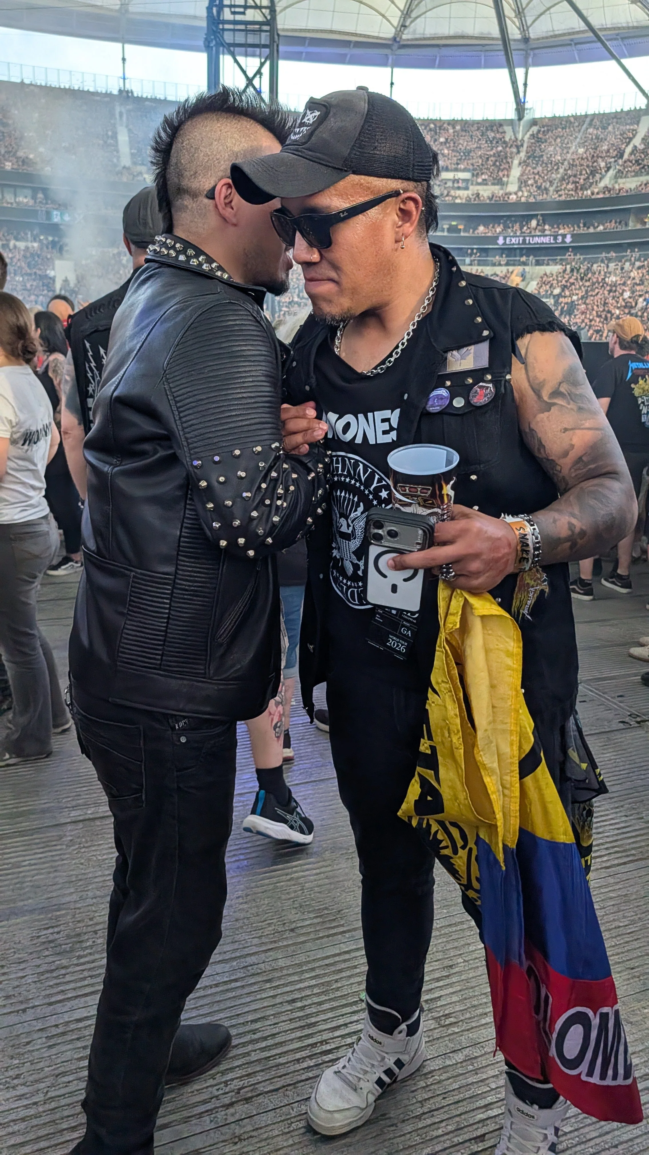 Two men with mohawk hairstyles and dark clothing are standing close together in a crowded stadium. One man is wearing sunglasses and a black cap, holding a cup and a phone, and has tattoos and jewelry. The other man is wearing a leather jacket with m