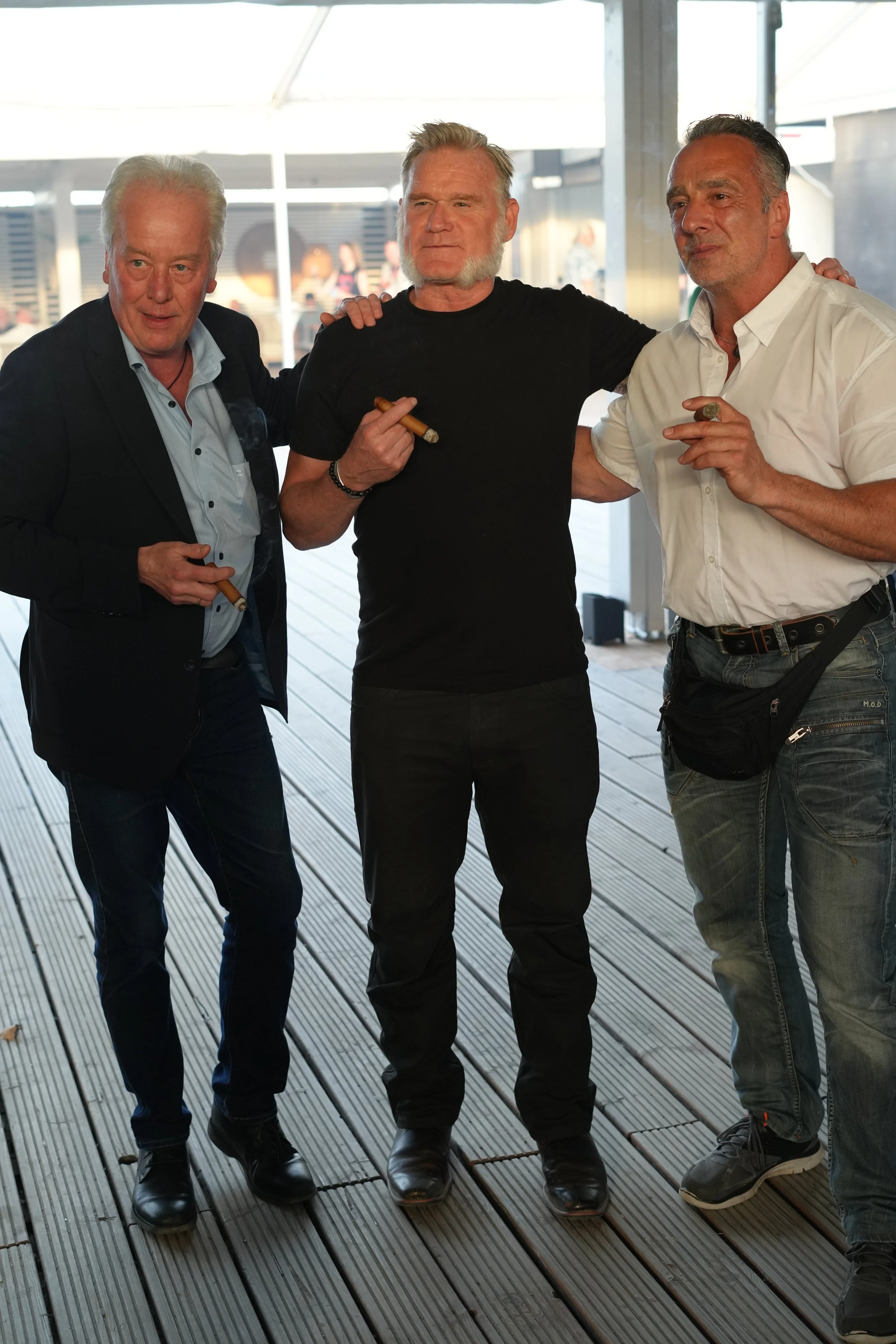 Three men standing close together, holding cigars, in a bright indoor space with a wooden floor and large windows.