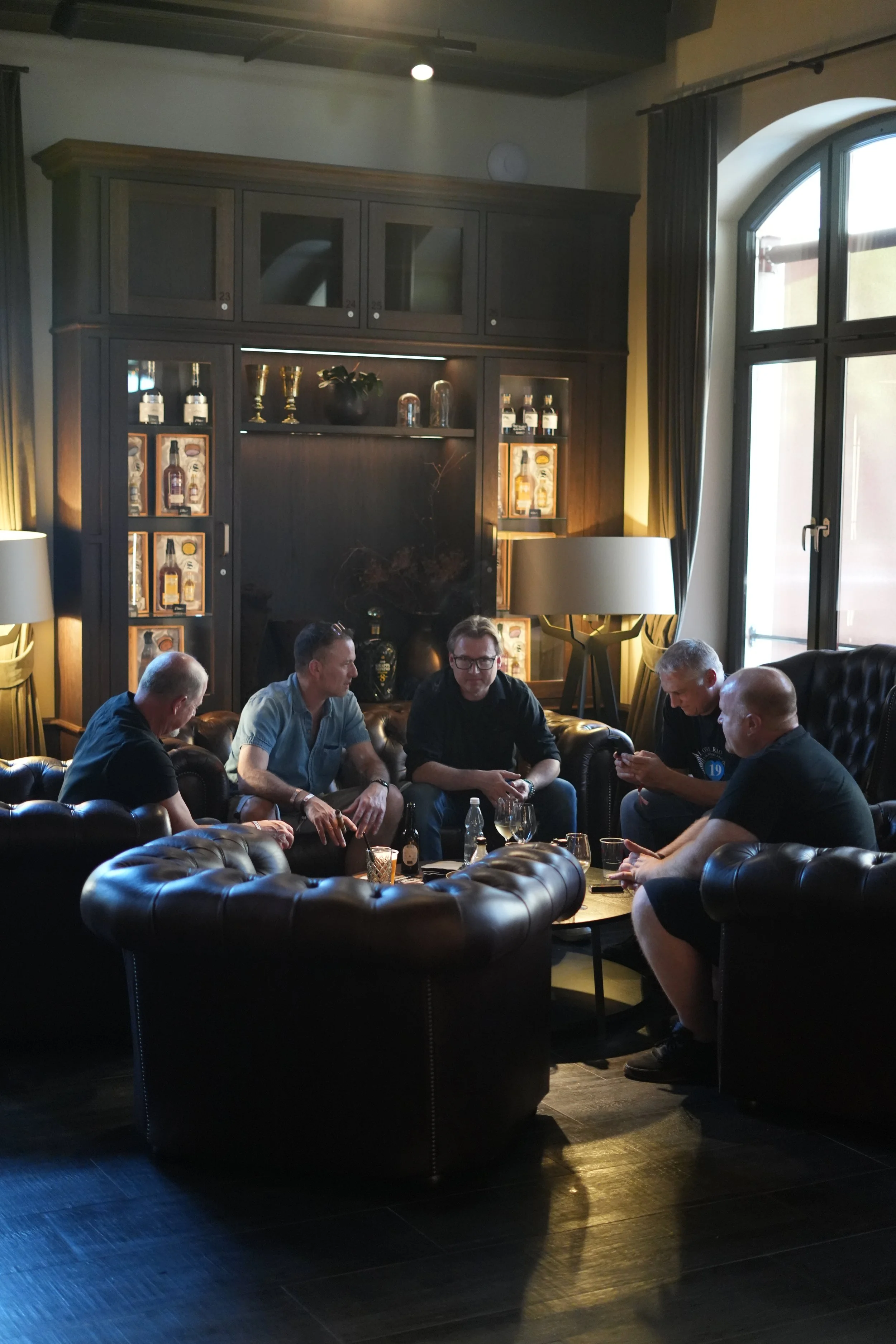 A group of five men sitting on leather sofas in a dimly lit living room or lounge area, socializing and using their phones. The room features dark wood furniture, large windows with curtains, and decorative items including bottles and trophies displa