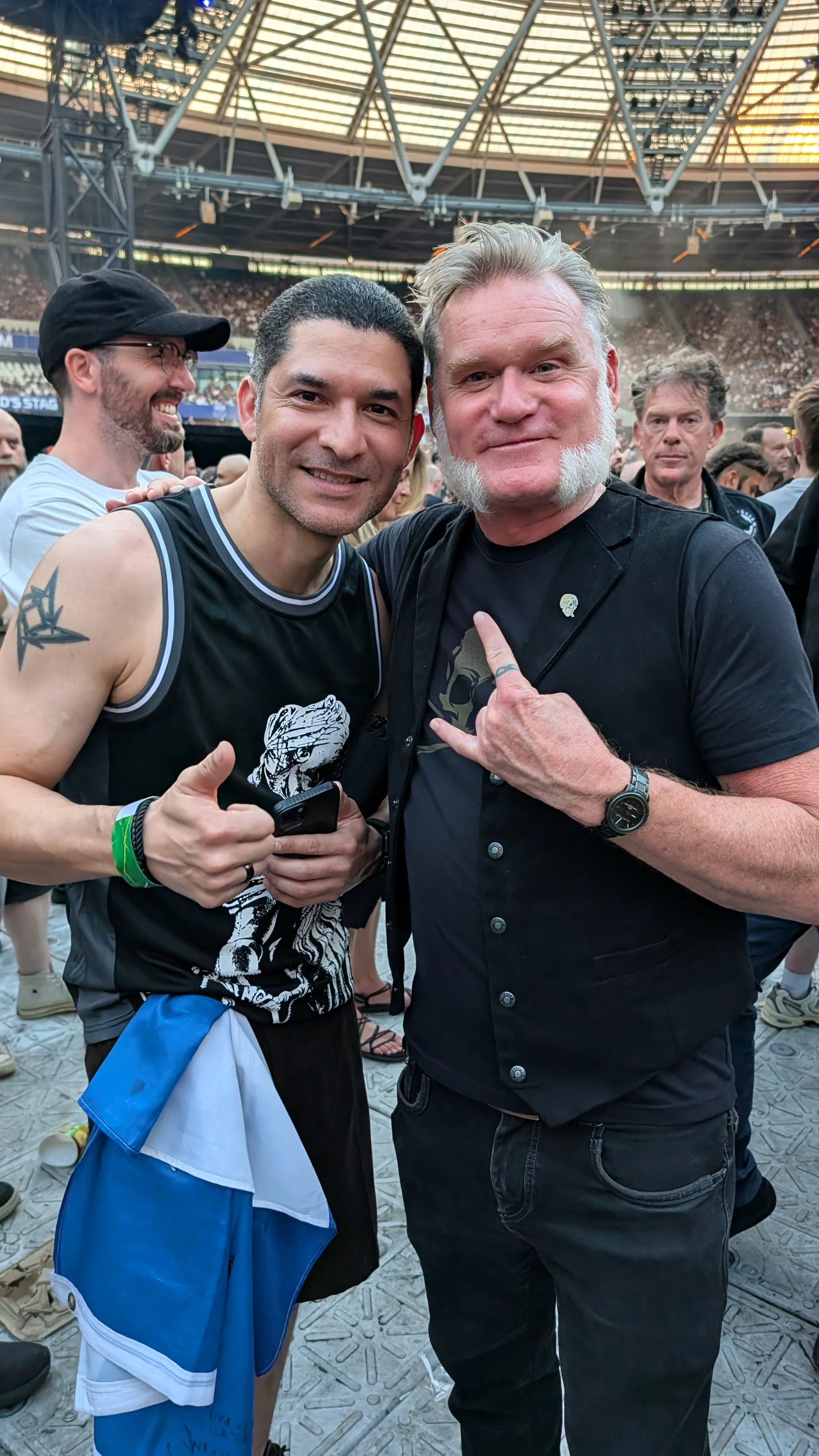 Two men taking a selfie at a crowded outdoor concert or event, with a large stage and many attendees in the background. Both are smiling and making gestures with their hands.