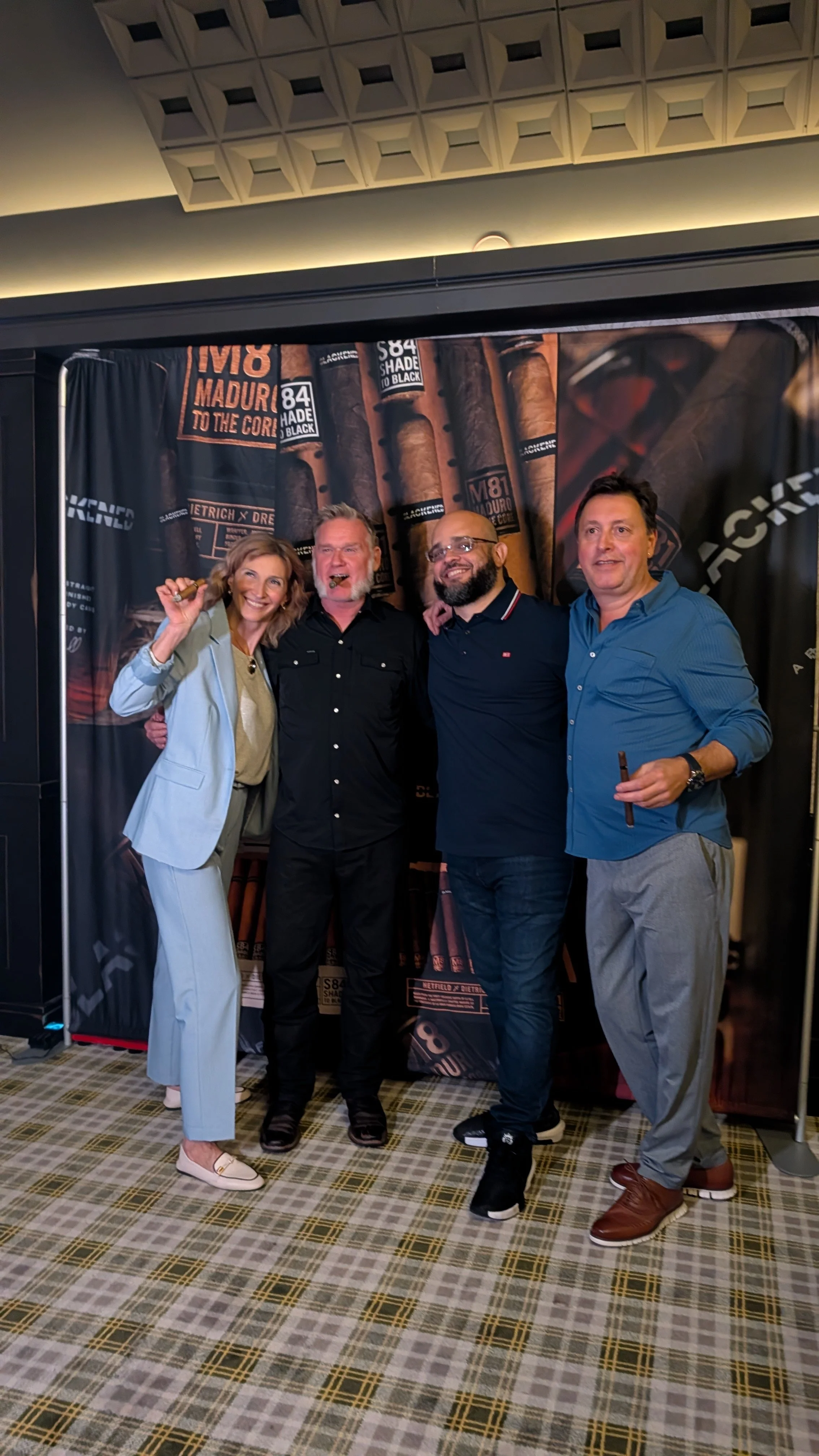 Four people standing together in front of a cigar-themed backdrop, smiling and posing for the photo. The group includes two men and two women, dressed in business casual attire.