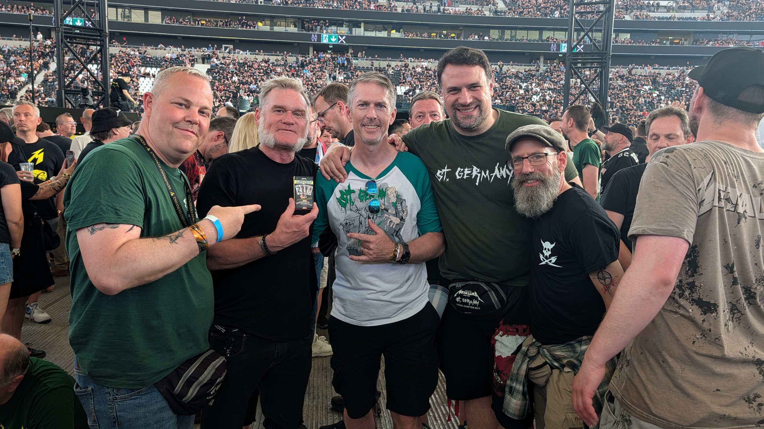 A group of five men at a concert or event, smiling and posing together in a crowded venue with a large audience in the background, some wearing band t-shirts.