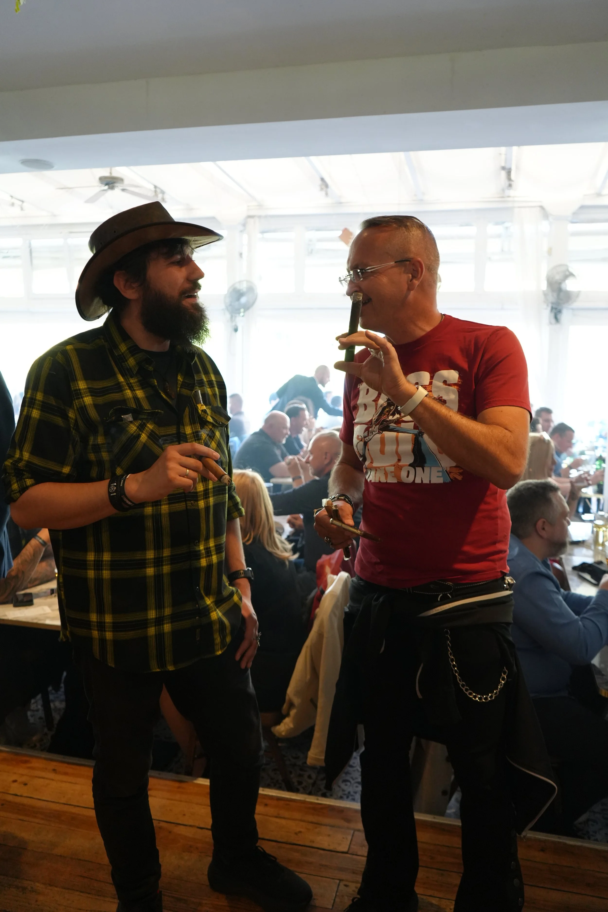 Two men are standing and engaging in conversation inside a busy restaurant or café. One man is wearing a brown cowboy hat and a yellow plaid shirt, and the other man is wearing glasses and a red graphic t-shirt. They are smiling and appear to be enjo