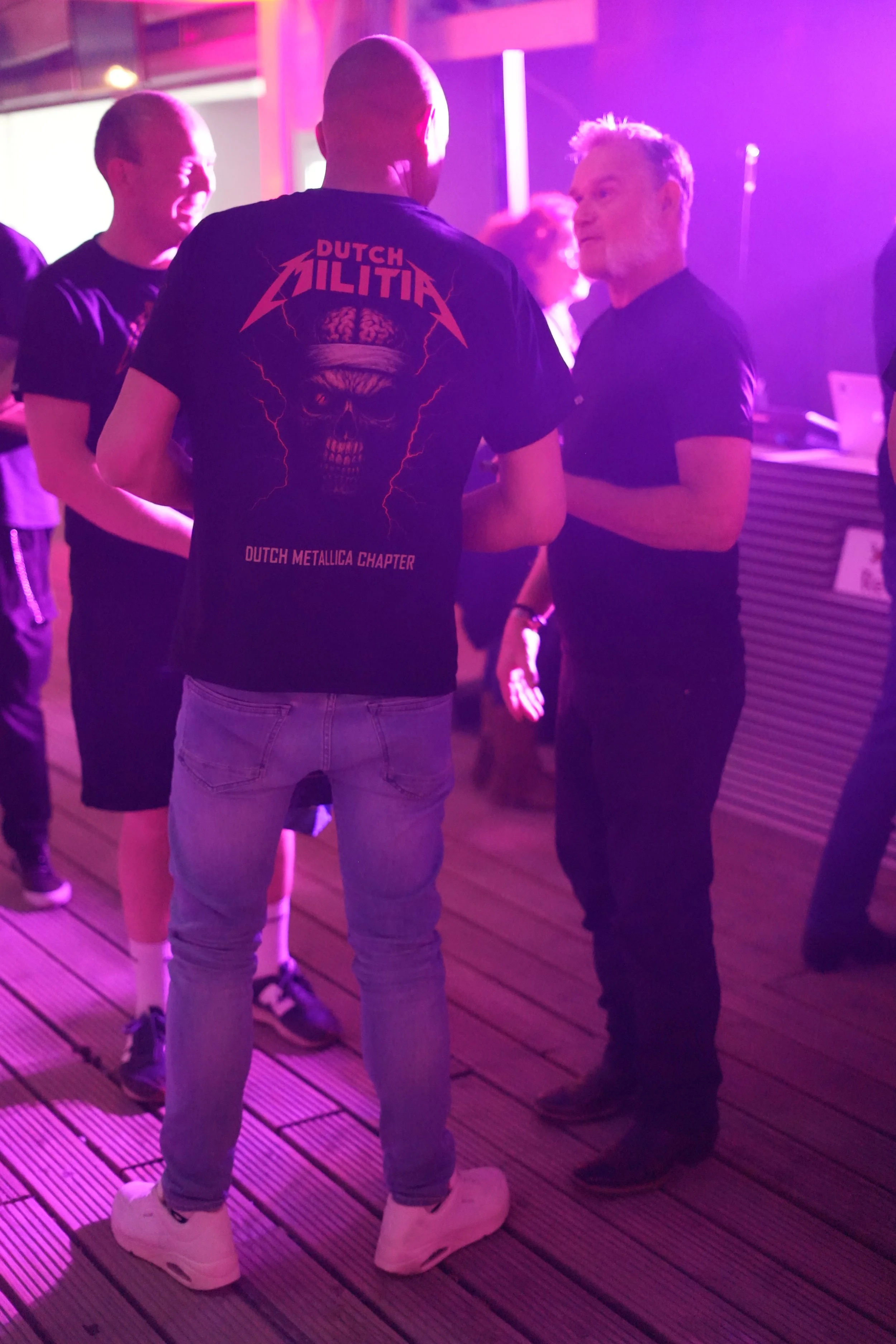 Three men are standing and talking in a dimly lit room with purple and pink lighting. One man has a shaved head and is wearing a black T-shirt with a design on the back featuring a skull and lightning bolts, with the words 'Dutch Metallica Chapter.' 