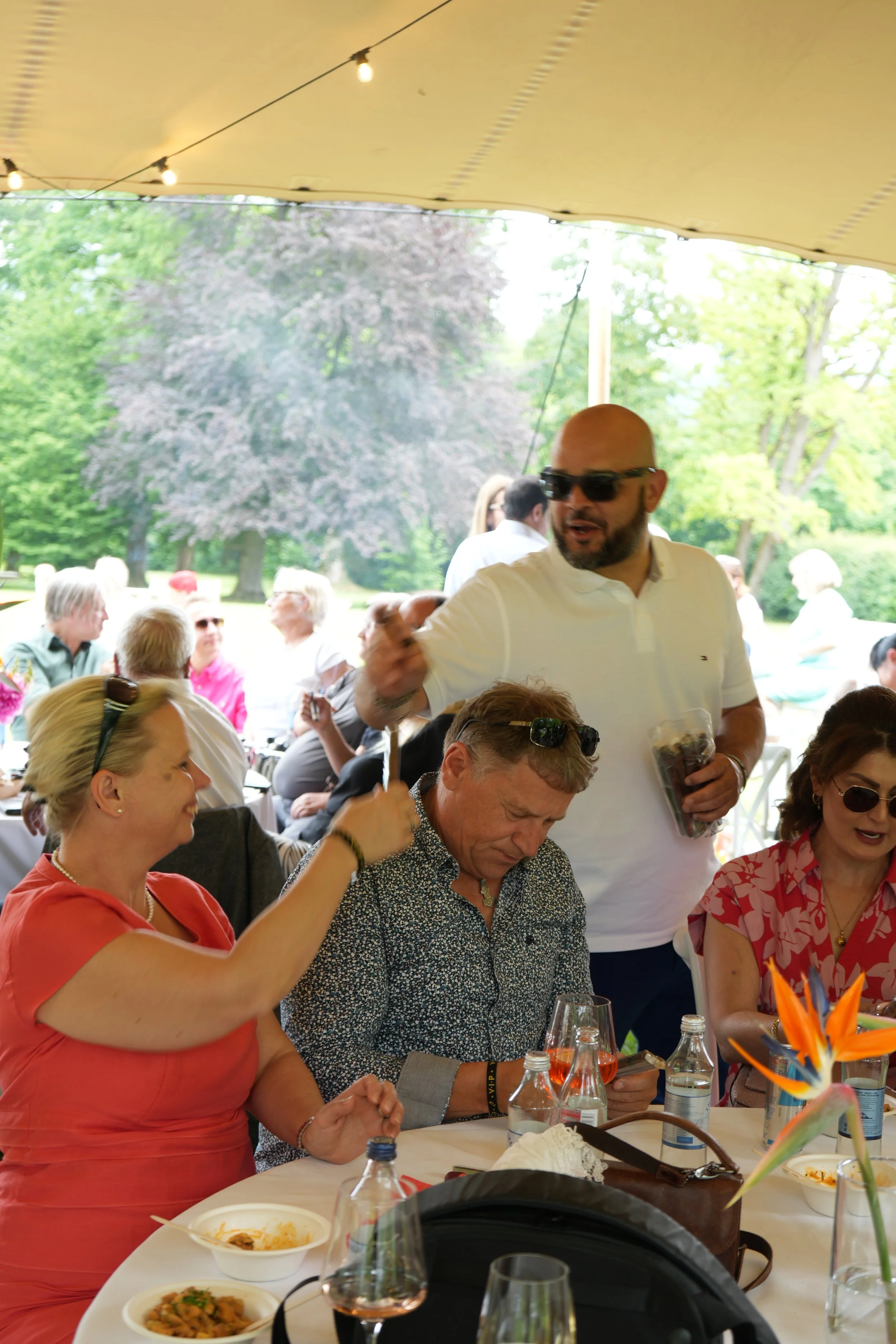 A group of people gathered around a table outdoors under a canopy with string lights, enjoying a meal.