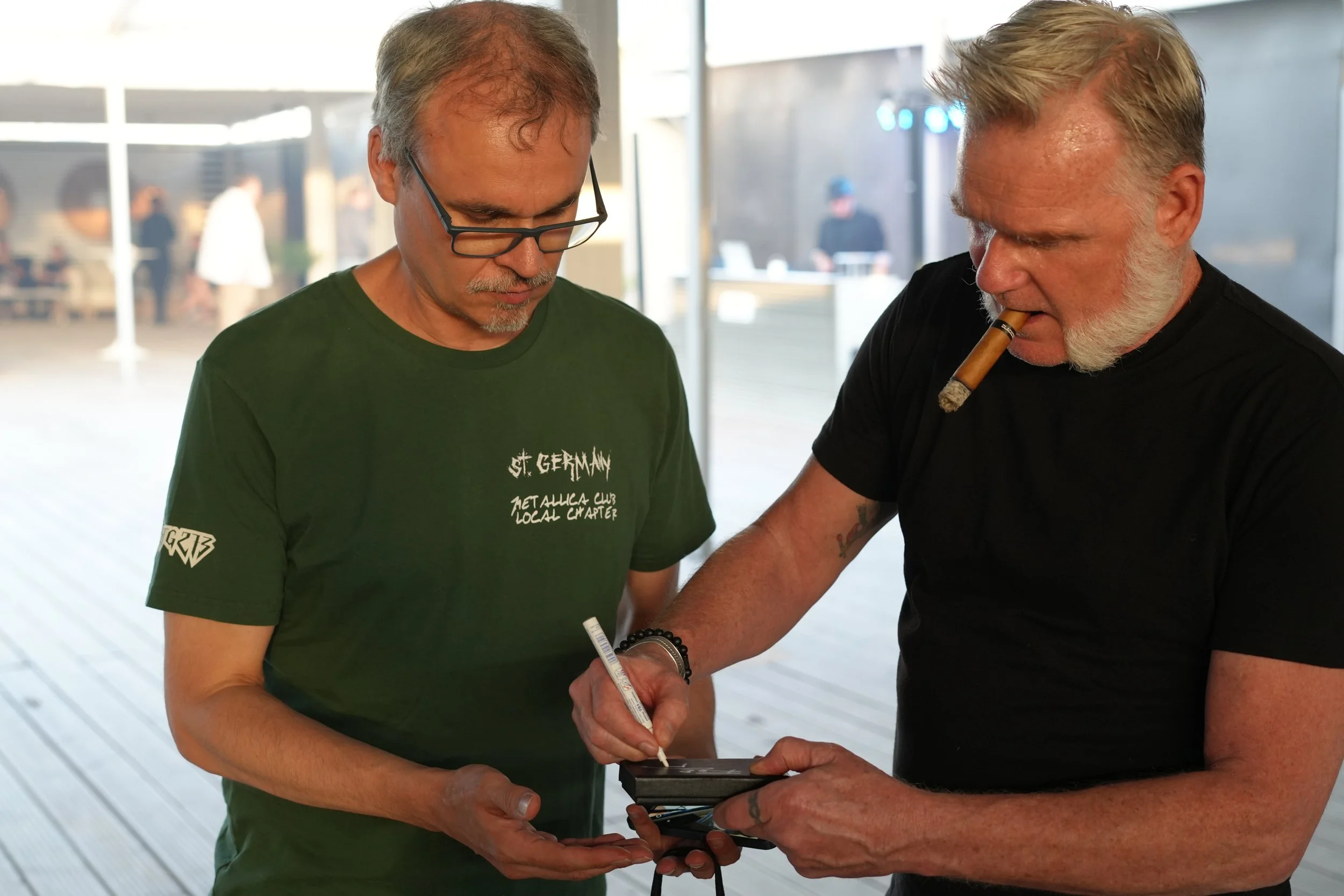 Two men are focused on an electronic device, one with glasses and a cigarette in his mouth, in a bright indoor setting with blurred people in the background.