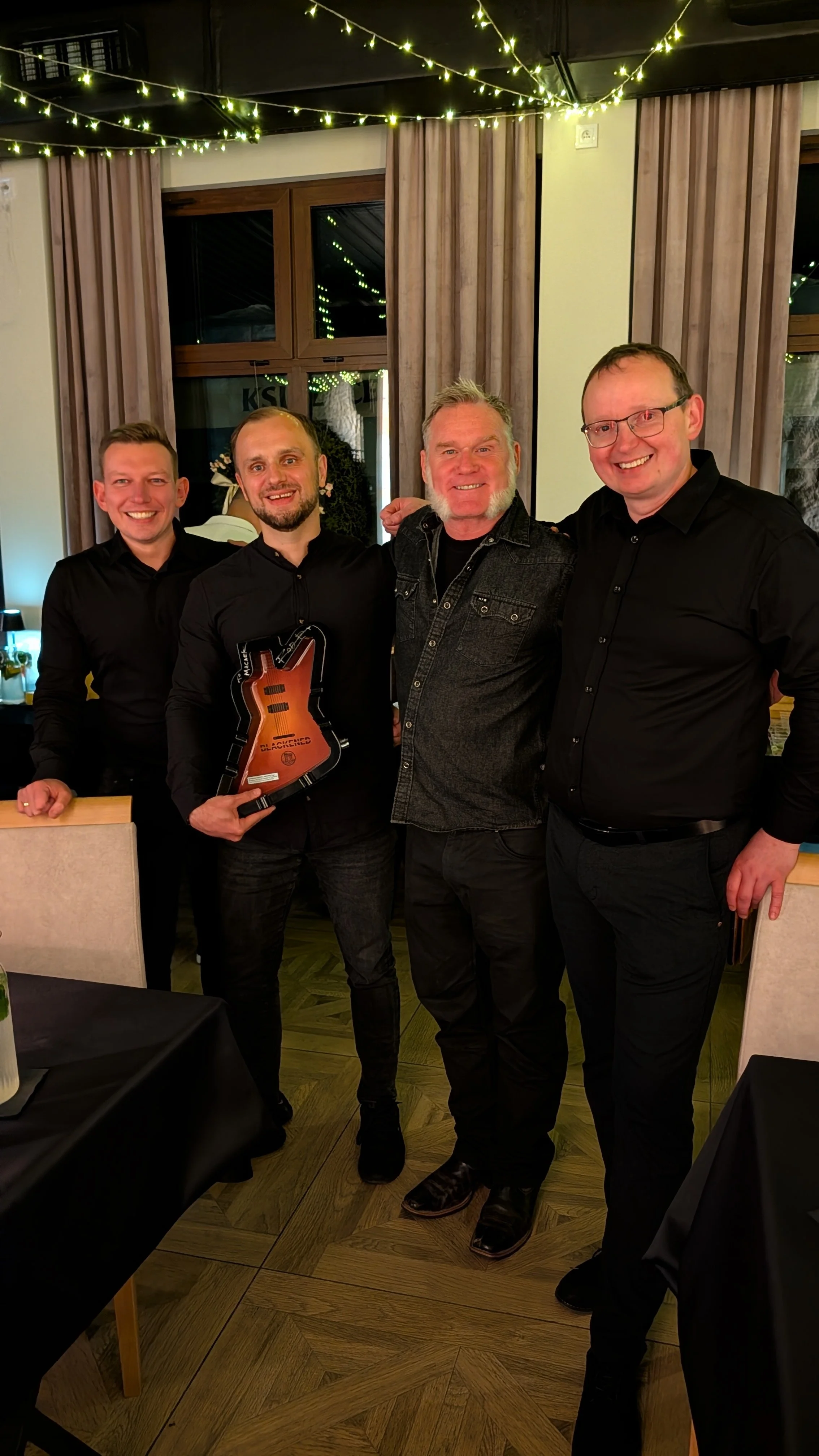 Four men standing together indoors, smiling, with one holding a guitar-shaped award, under string lights and wooden window frames.