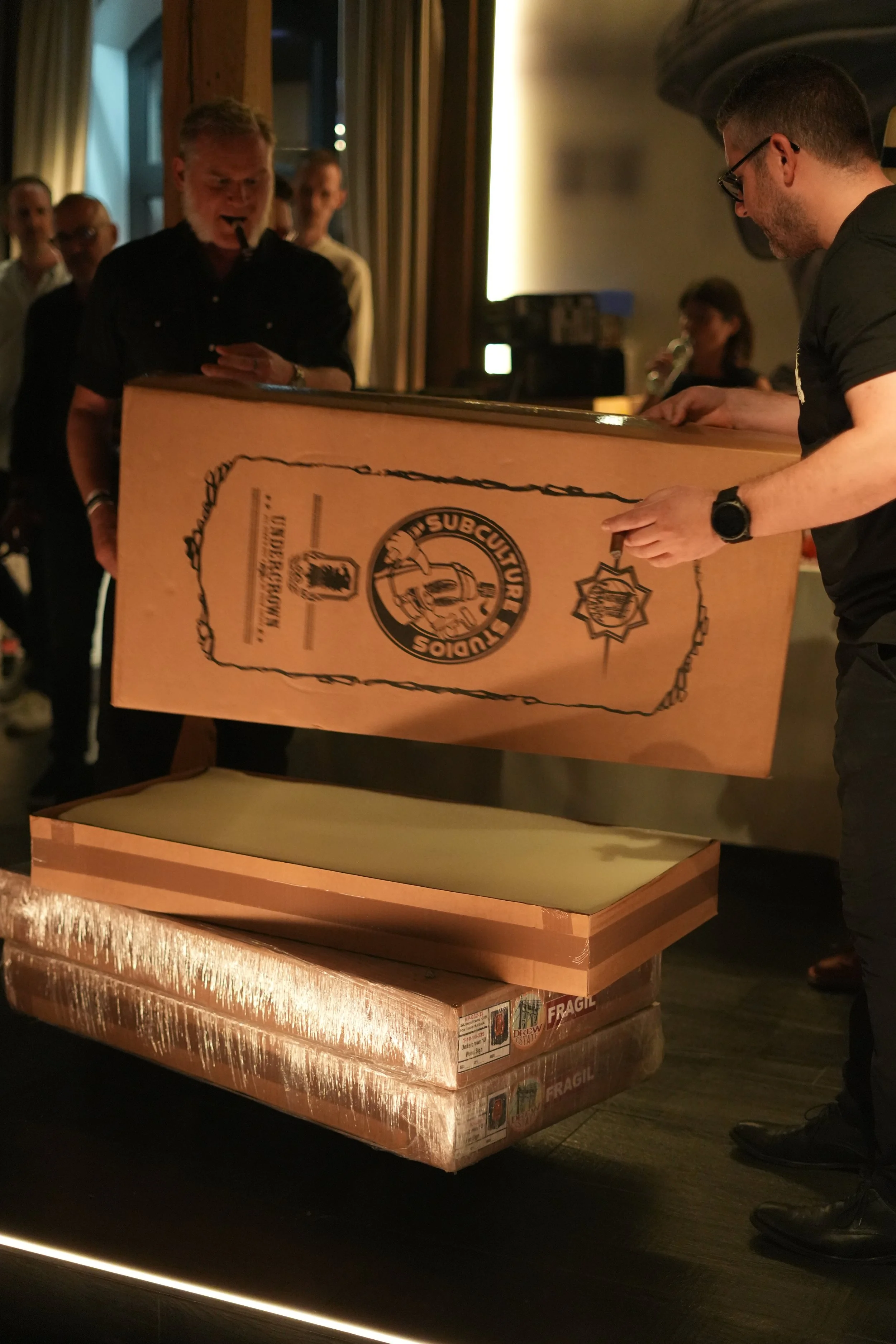 A man is lifting a large wooden box with a logo and text on it, while other people observe. The scene appears to be indoors, possibly in a store or event space.