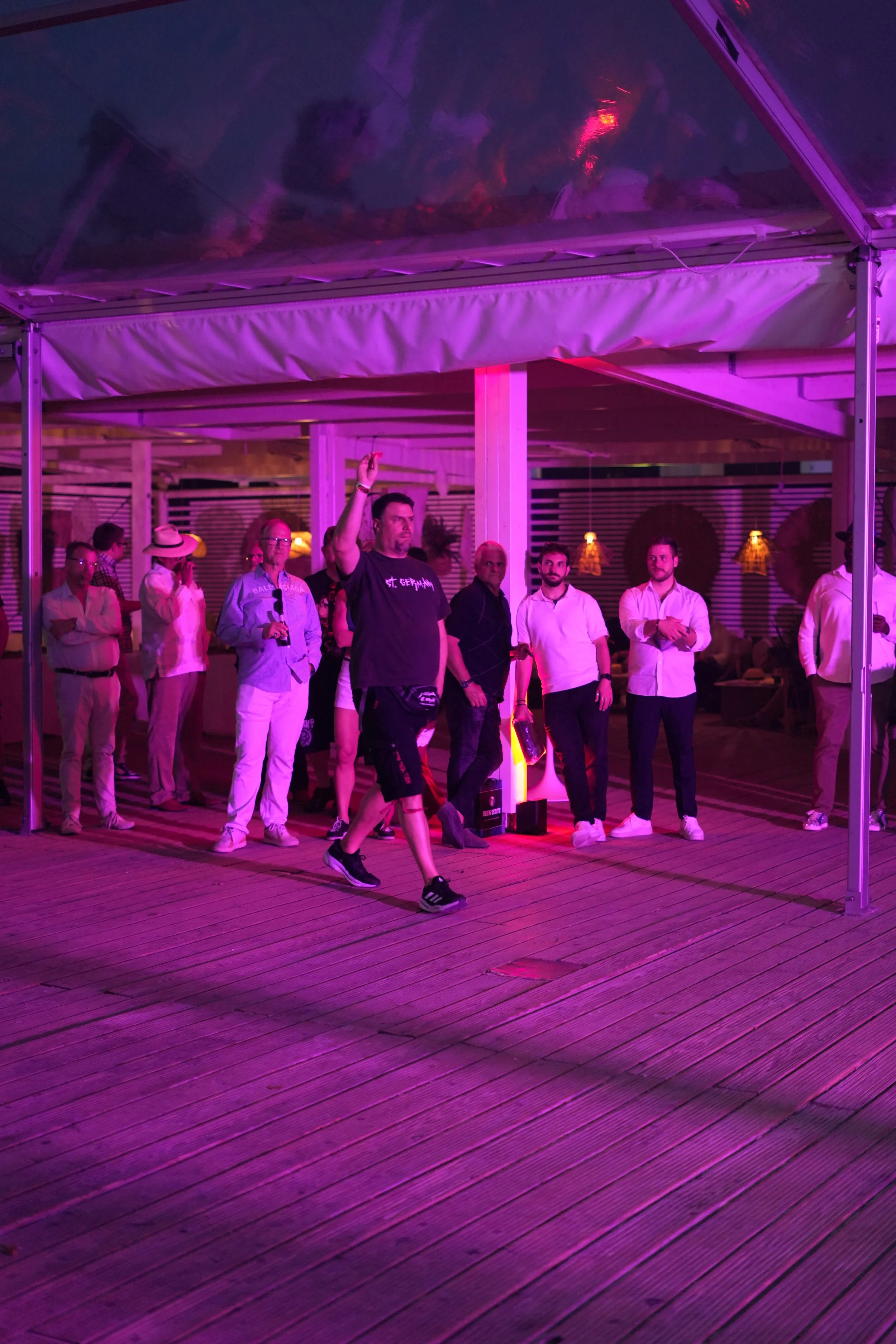 People standing under a pink-lit canopy at night, some engaged in conversation, with a man walking in front holding a bottle.
