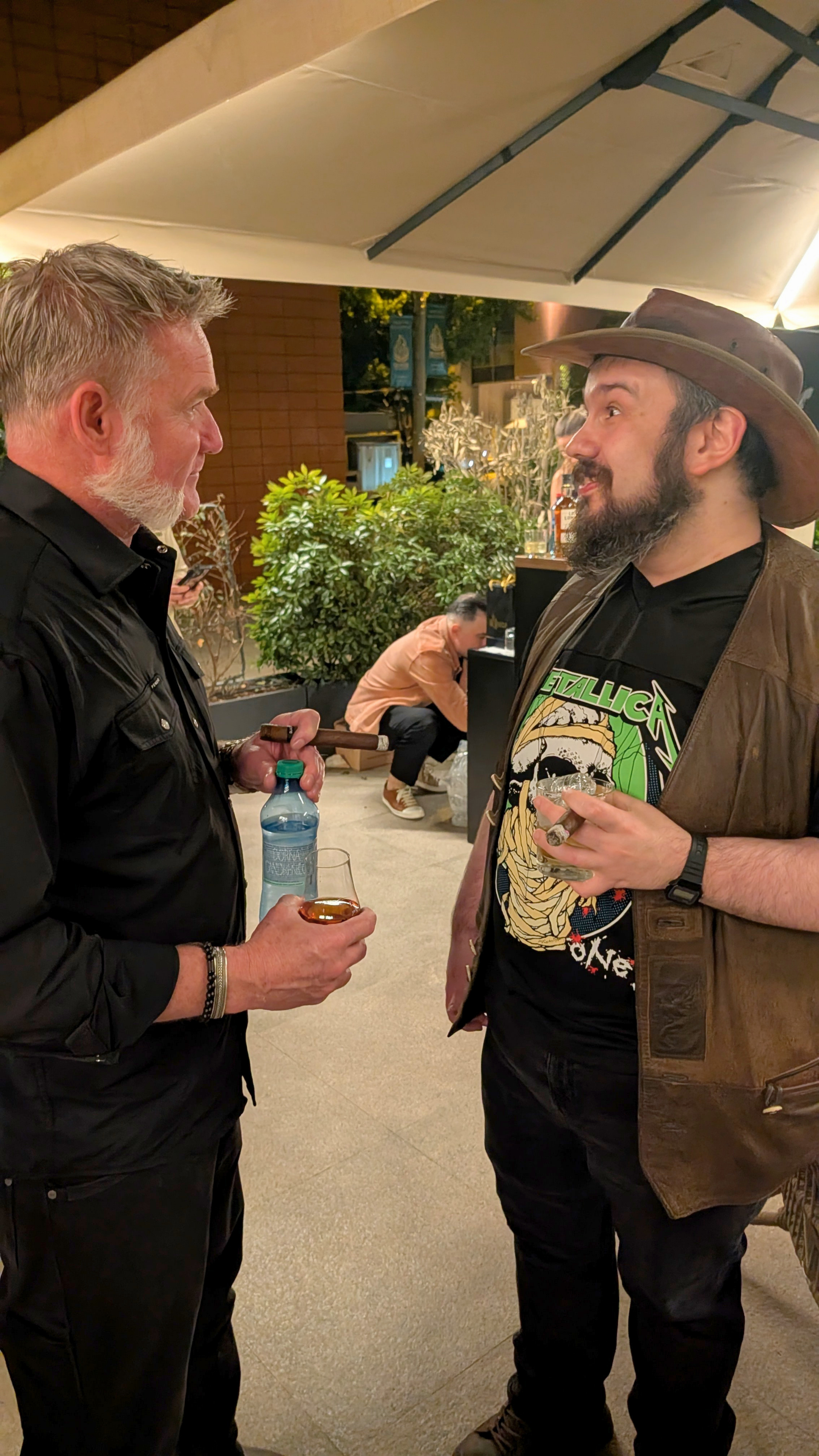 Two men are conversing at an outdoor social event. One man has gray hair and a beard, and the other has a beard and is wearing a hat. They are holding drinks and are dressed casually, with one man wearing a Metallica t-shirt.