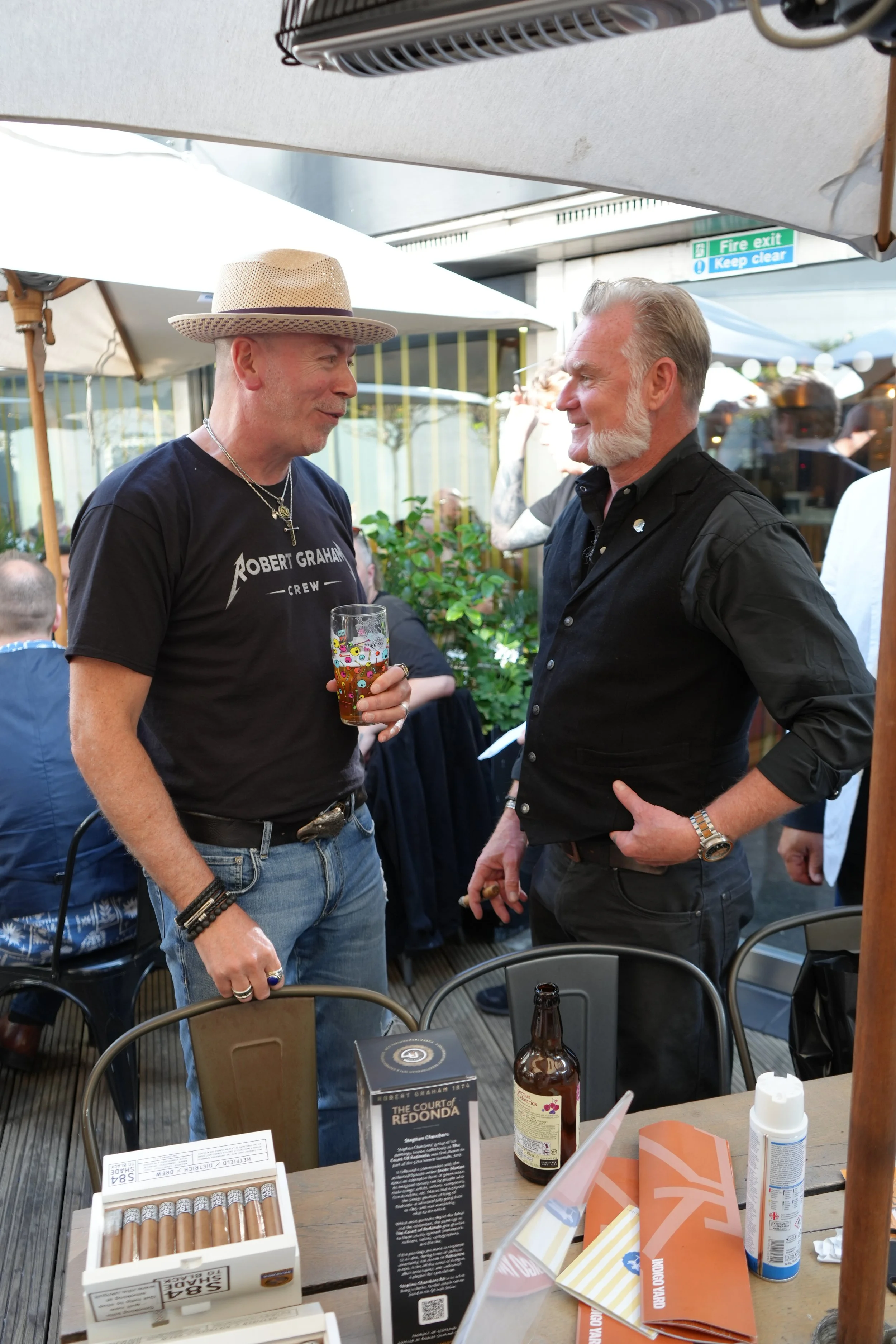Two men are engaged in a conversation at an outdoor event. One is wearing a straw hat, a Robert Graham T-shirt, and has a glass of beer in his hand. The other is dressed in a black shirt with gray hair and beard, and is smiling. There are tables with