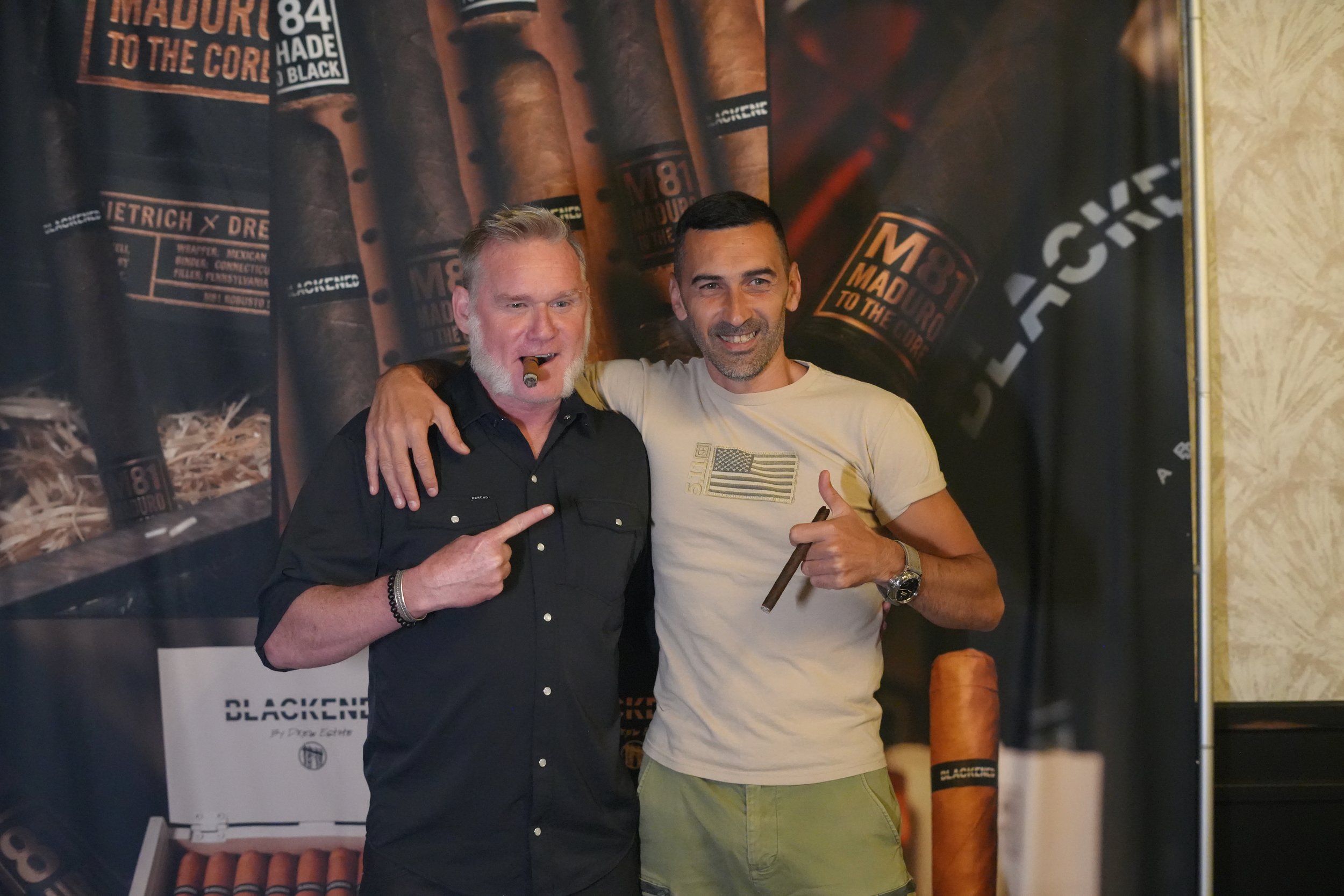Two men standing together, smiling and posing with cigars, in front of a backdrop with cigar images and branding for Blackened by Dee Snider.
