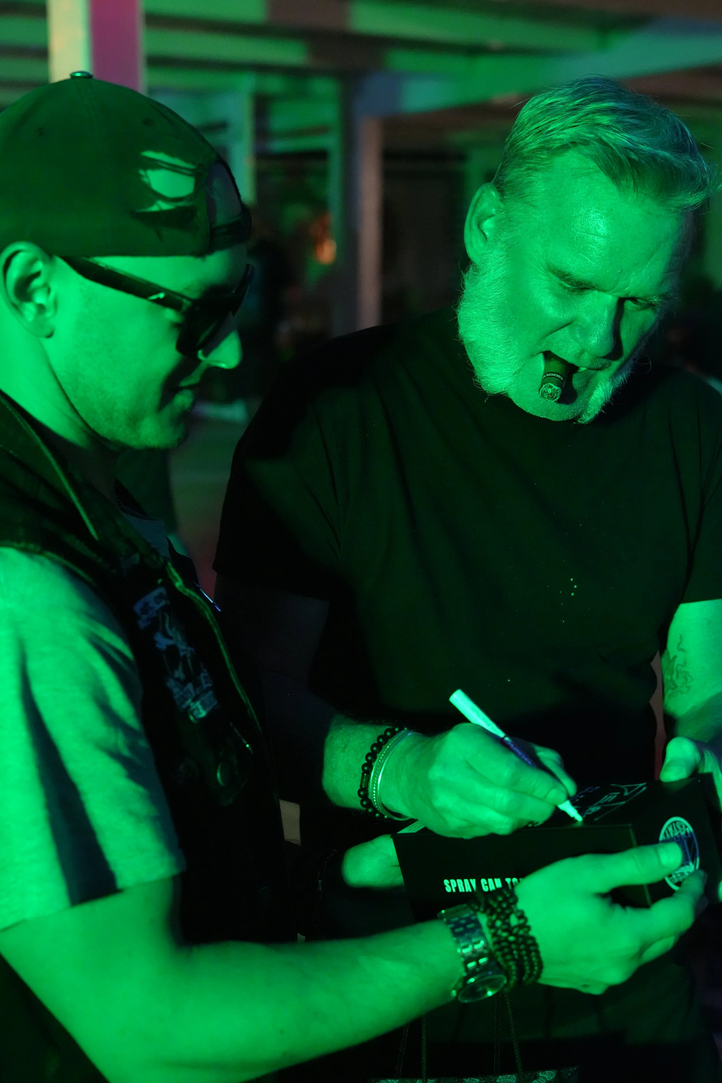 Two men are signing autographs under green lighting, with one holding a black box that reads "SPRAY CAN TO".
