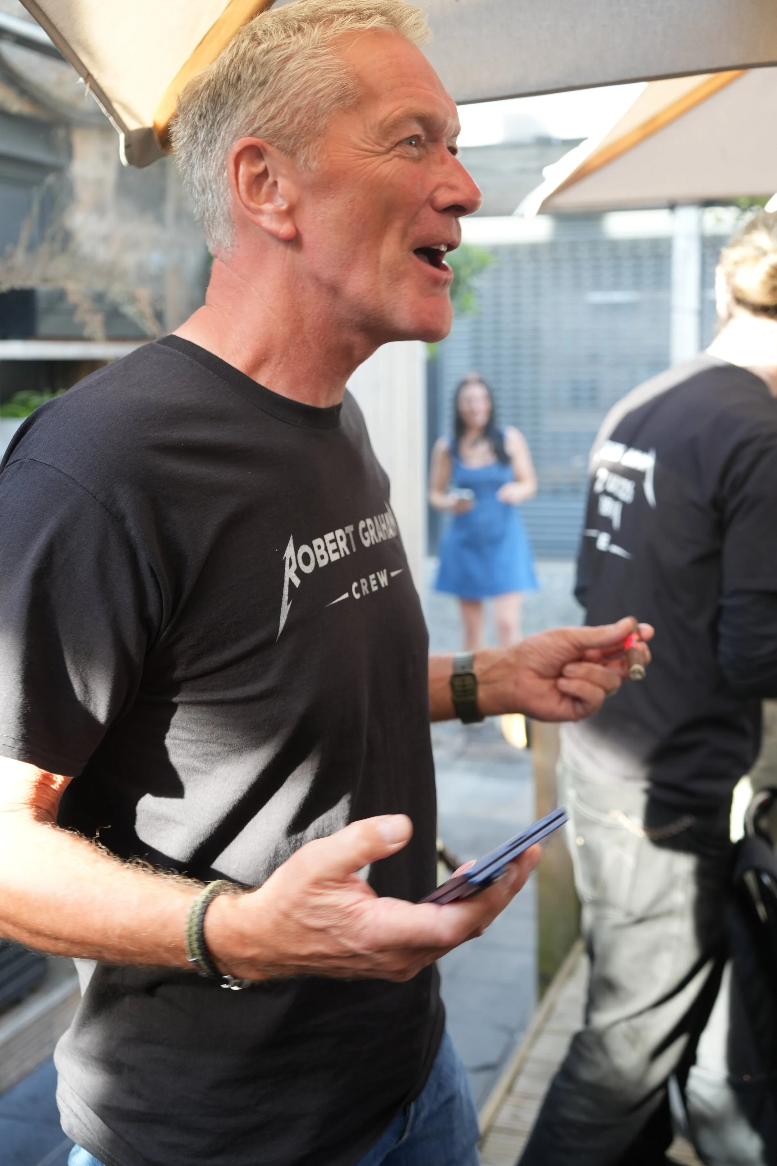 A man with gray hair wearing a black t-shirt holding a smartphone and speaking at an outdoor gathering.