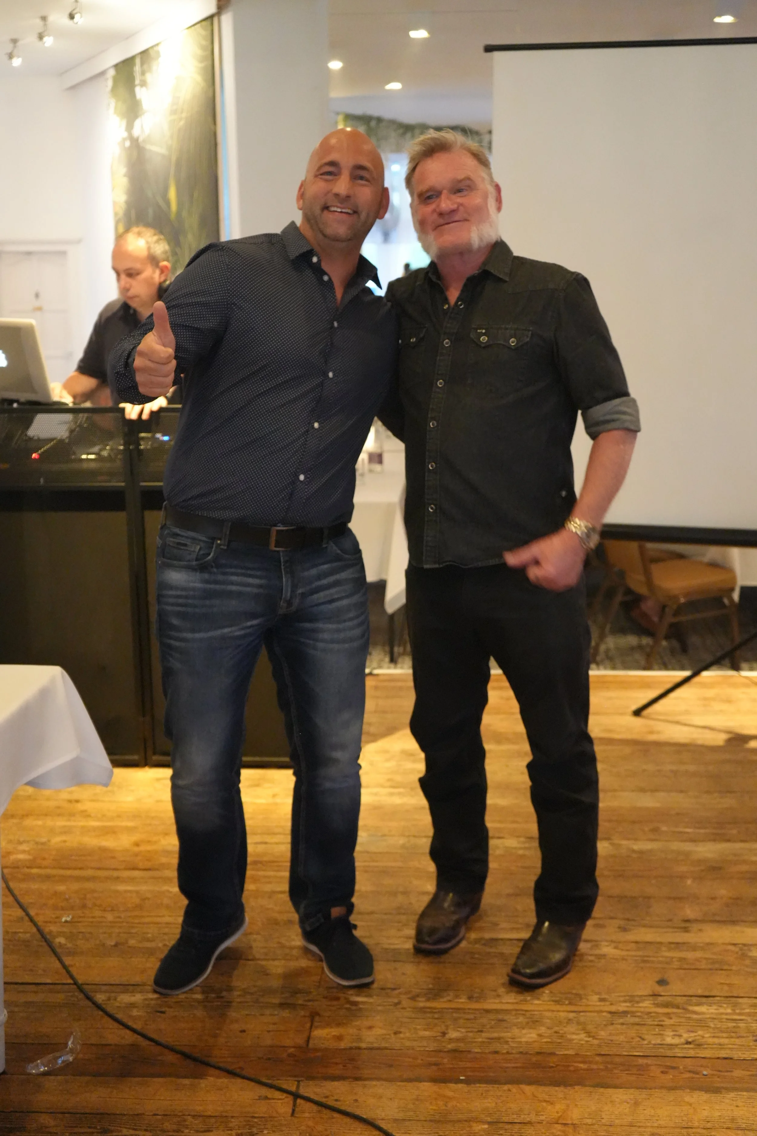 Two men standing close together, smiling, one giving a thumbs-up, inside a room with wooden floor, white walls, and a large white projection screen. A DJ is seen in the background working on a laptop.