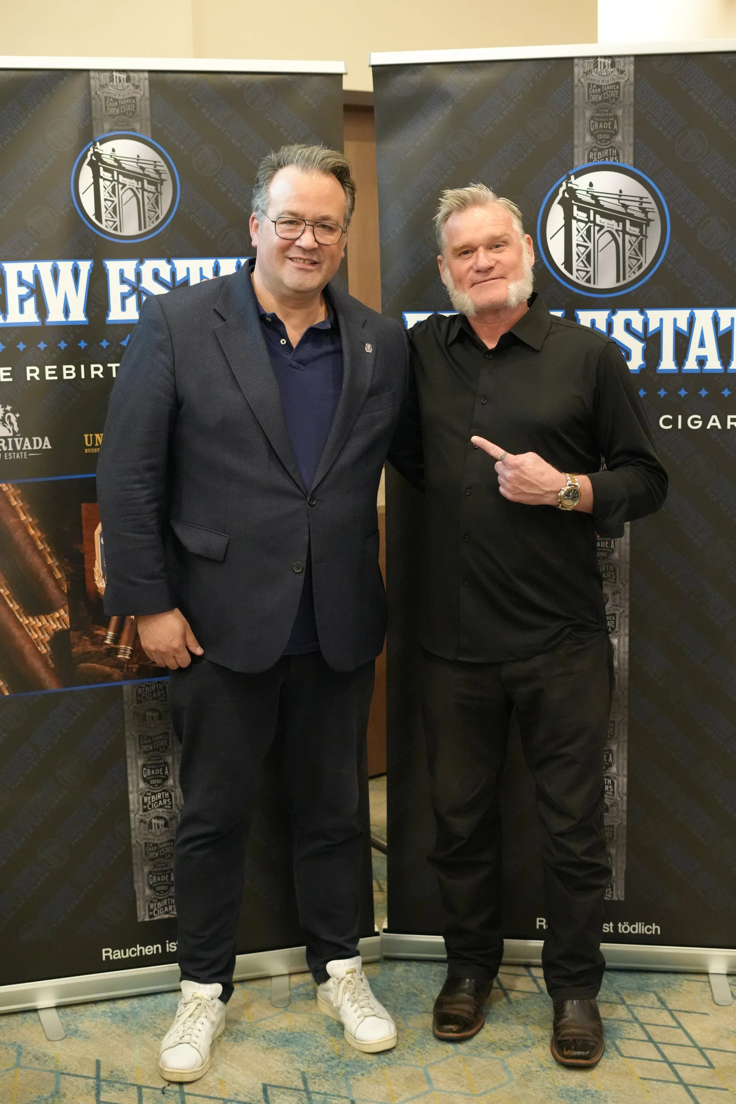 Two men standing in front of banners with a bridge illustration and text. The man on the left is wearing glasses, a dark blazer, a navy blue shirt, and white sneakers. The man on the right has white hair, a beard, and is wearing a black shirt, black 