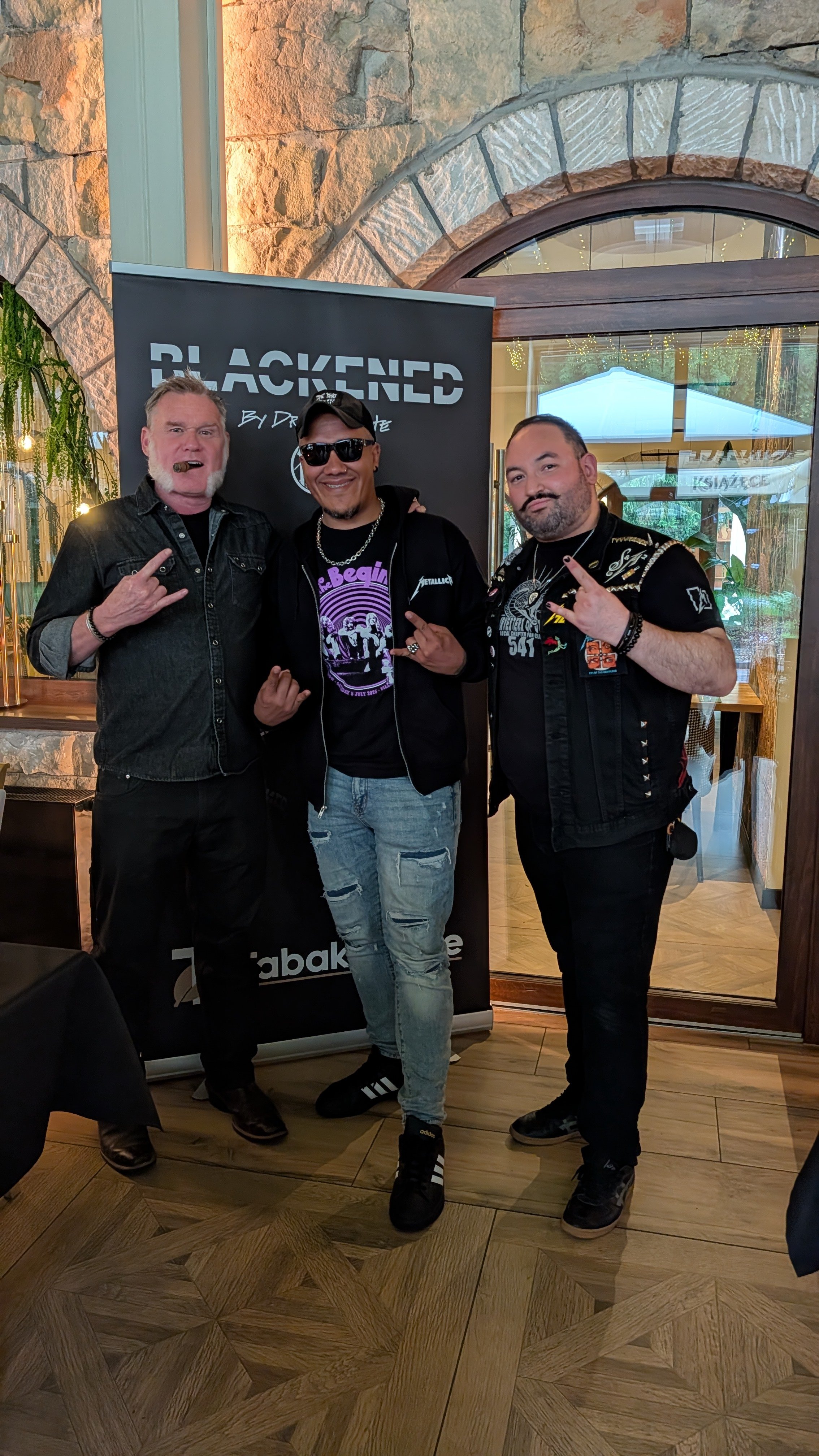 Three men posing in front of a black banner with the word 'BLACKENED' in large letters inside a restaurant or bar with stone walls and wooden flooring. All three are making hand signs and smiling or making expressive faces.