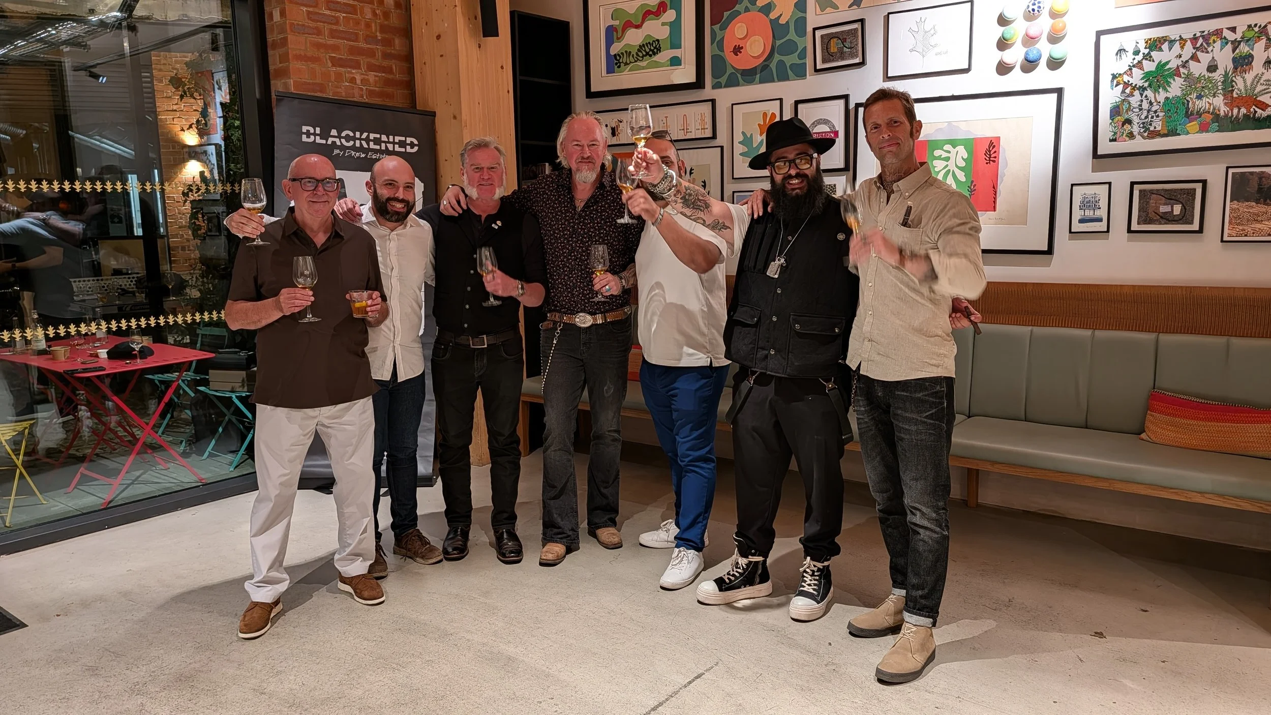 Group of eight men standing together inside an art gallery or restaurant, raising glasses in a toast, with colorful artwork on the wall behind them.
