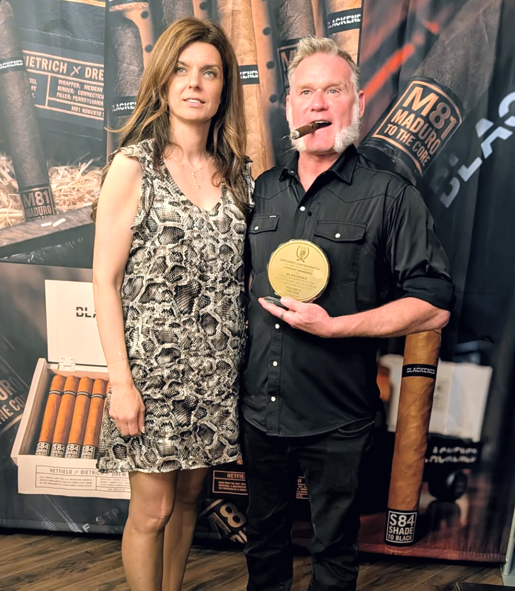 A man and woman standing together at an award event, the man holding a cigar in his mouth and a gold medal. The background features cigar branding and packaging, with the man wearing a black shirt and the woman in a leopard print dress.