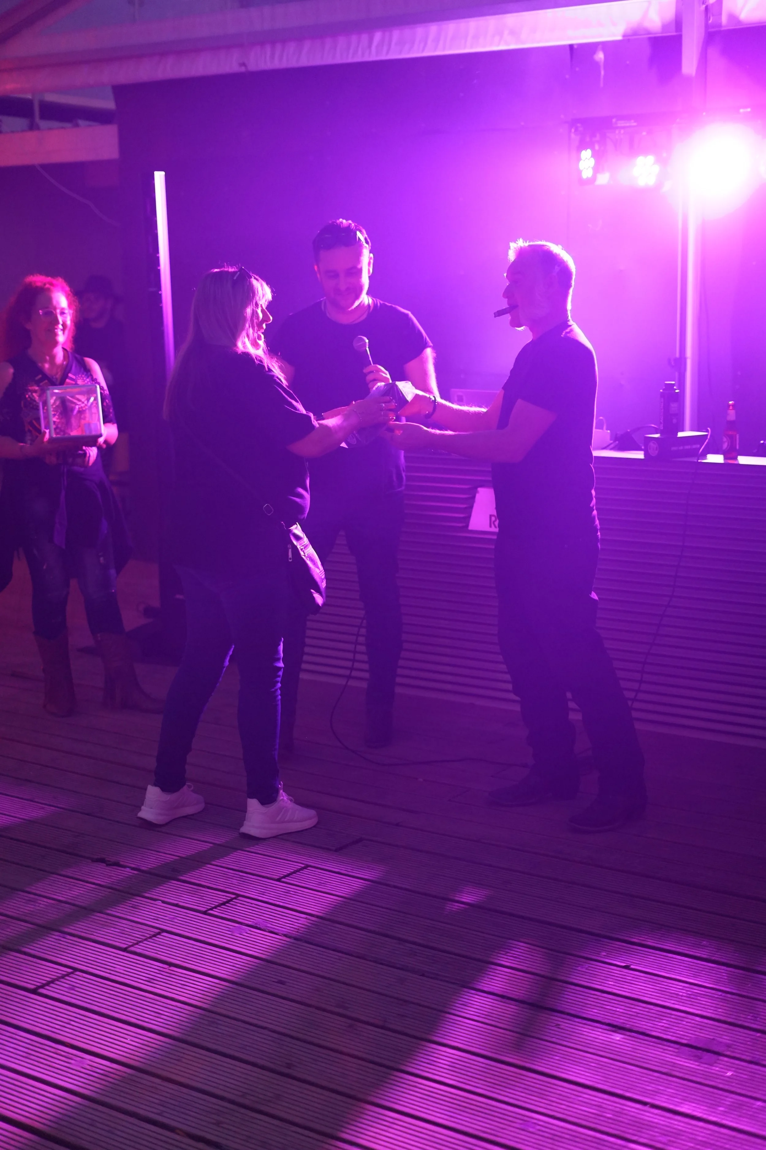 Group of people at a party or event, with one person handing something to another, illuminated by purple lighting, with a DJ booth in the background.