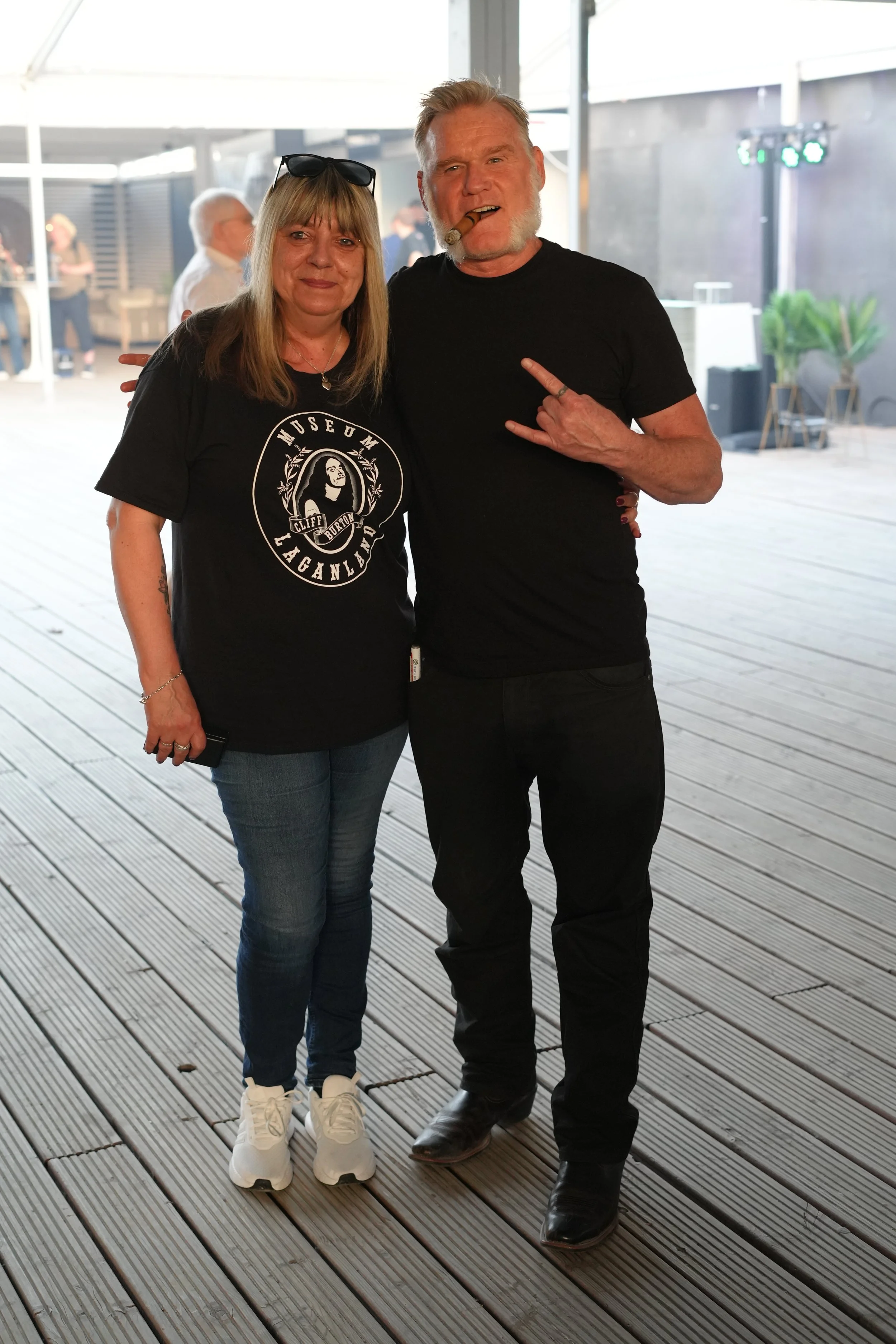 A man and woman standing together indoors, with the man smoking a cigar and the woman smiling, both wearing black shirts. The woman has sunglasses on her head and the man is making a rock sign with his hand.