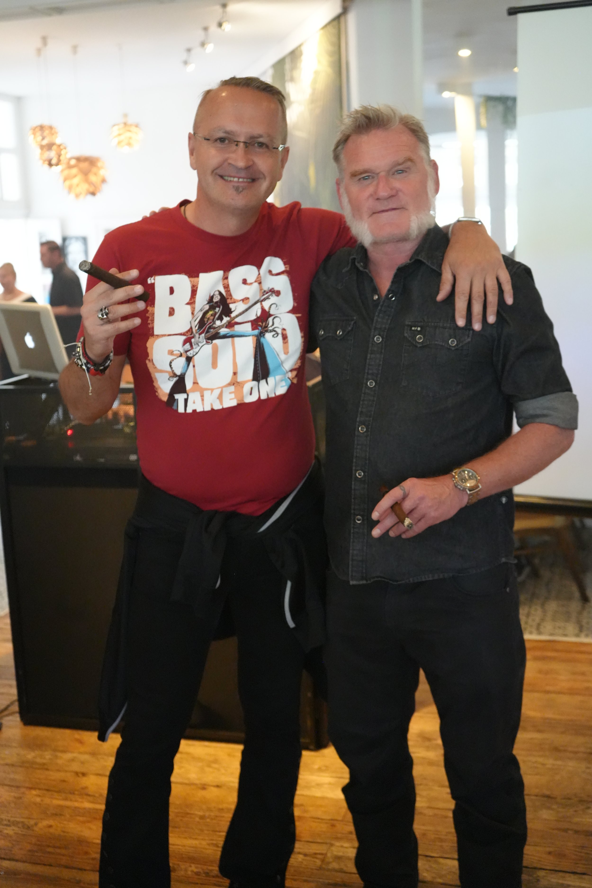 Two men standing arm-in-arm in a social gathering. The man on the left is wearing glasses, a red graphic T-shirt, and black pants, holding a cigar. The man on the right has a gray beard, is wearing a black denim shirt, and a watch. They are smiling a