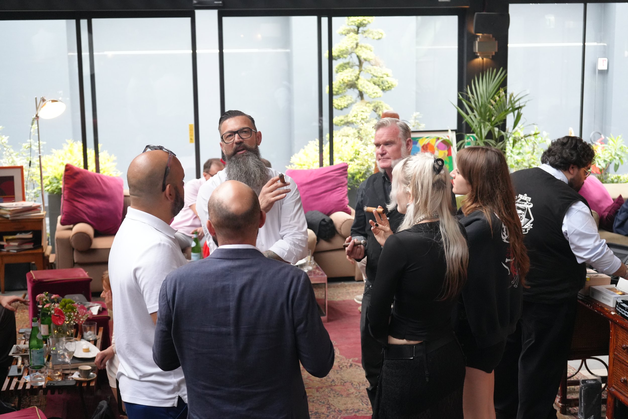 People socializing at a gathering inside a modern room with large glass windows, plants, and a table with drinks and snacks.