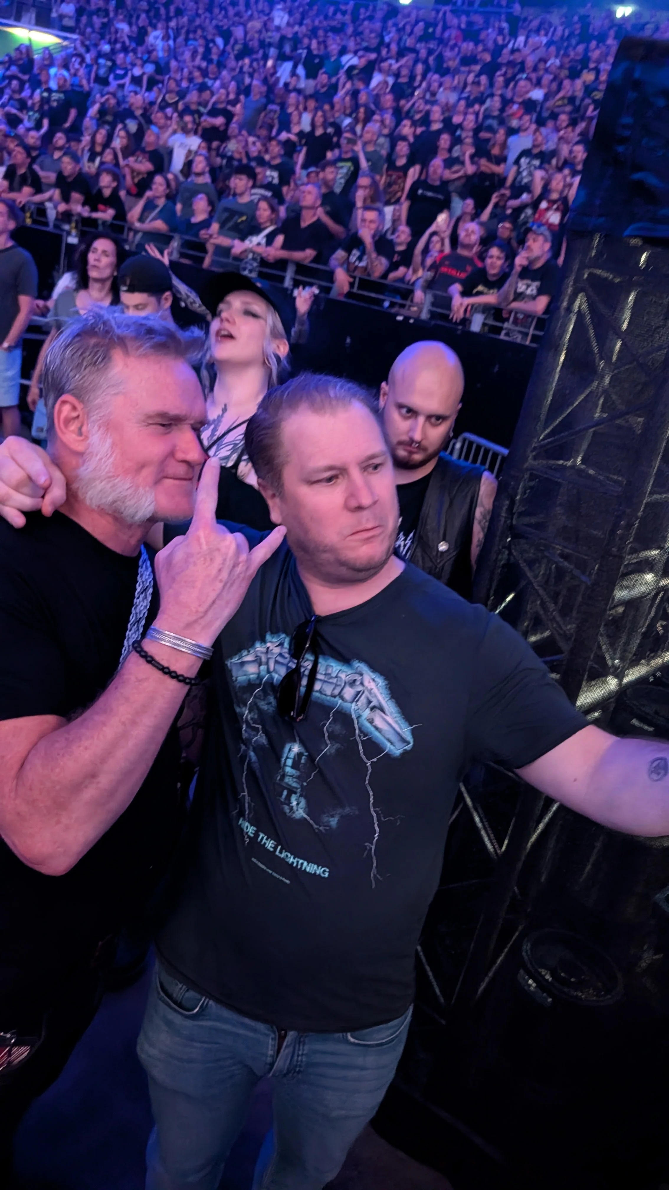 A man with gray hair and a beard making a rock on hand sign in front of a large crowd at a concert or music event.