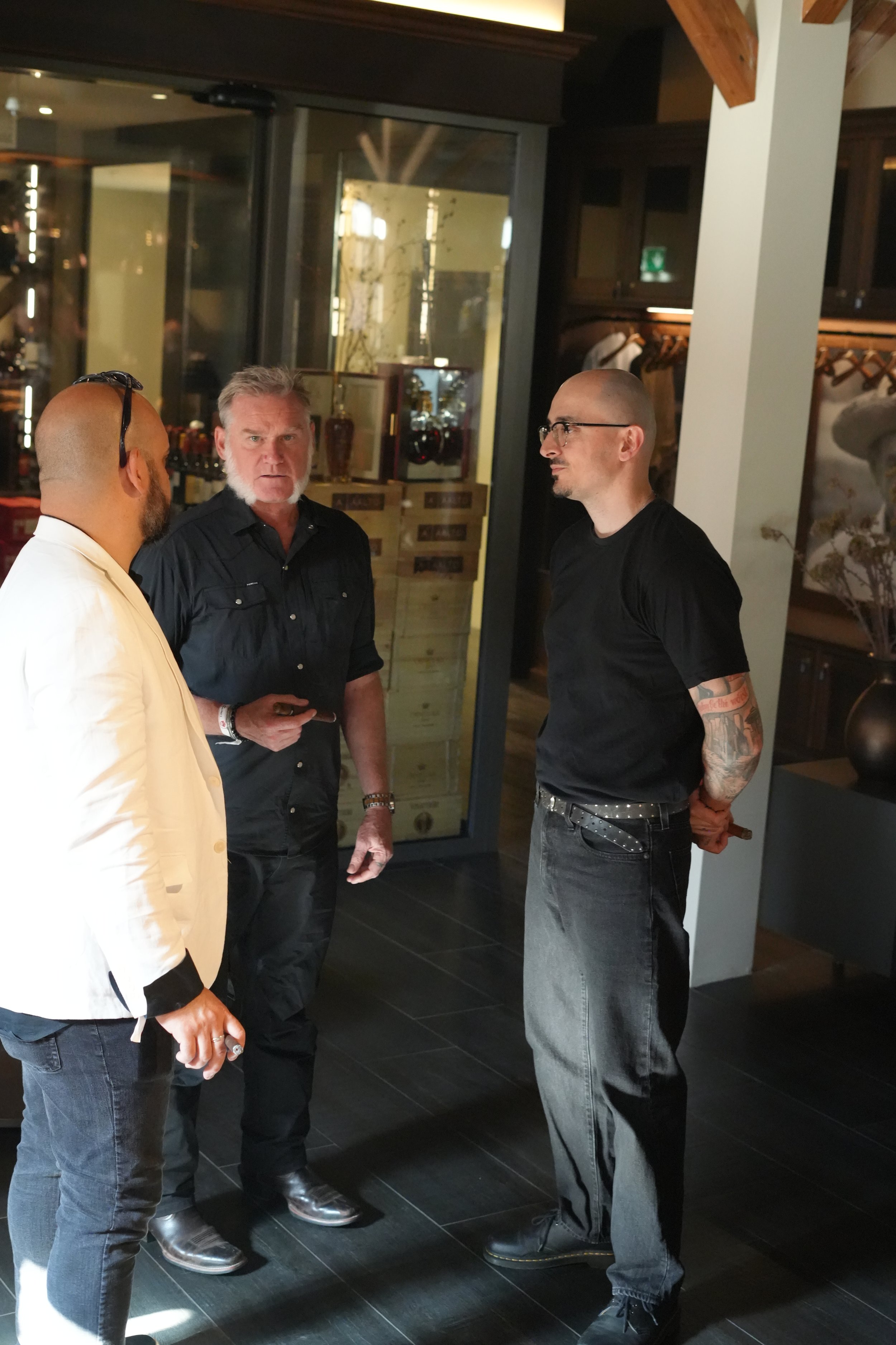 Four men are standing in a restaurant or bar, engaged in conversation. The men are dressed casually, with one wearing a white blazer, two in dark shirts, and one in a black T-shirt with tattoos on his arm. The background features dark wooden decor, g